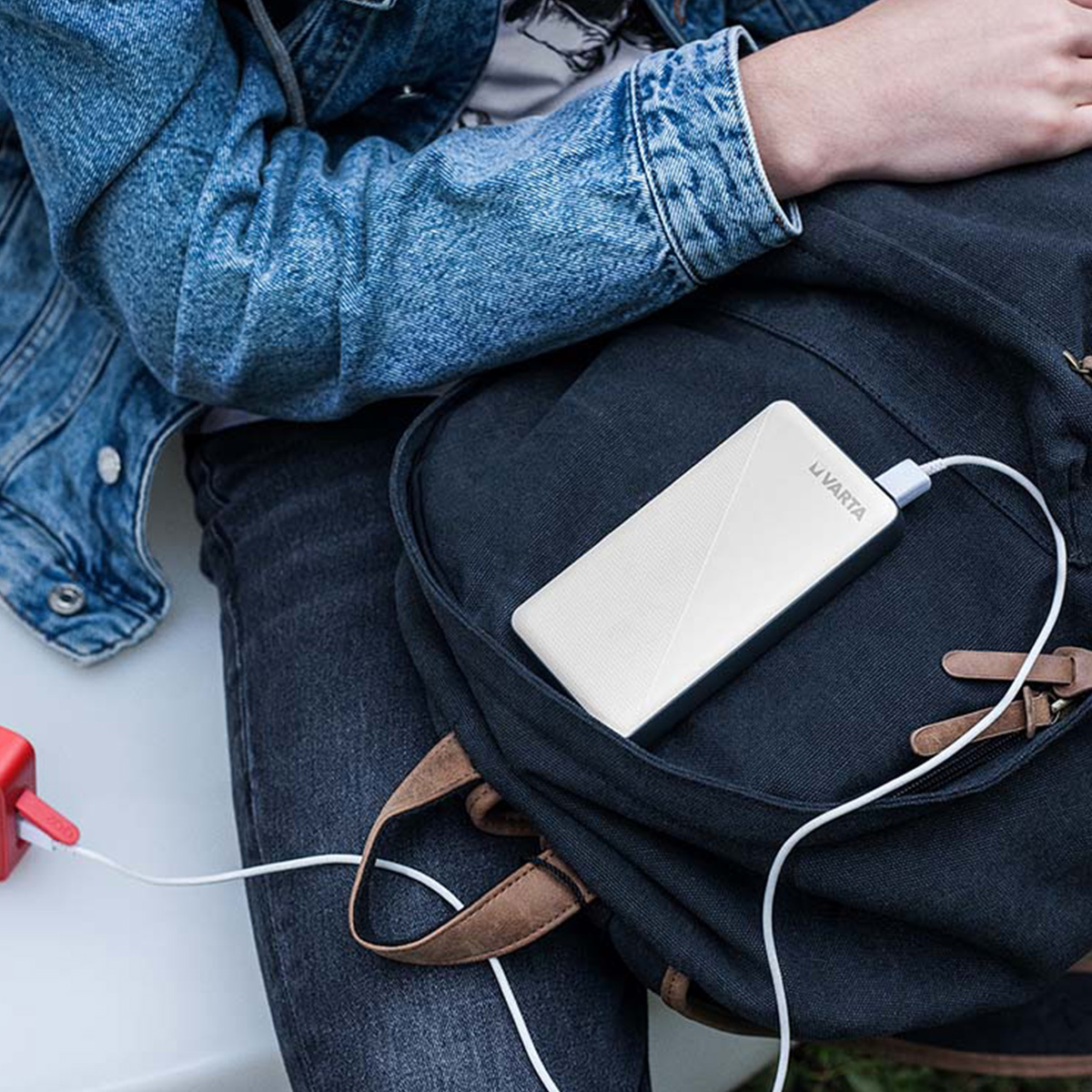 Eine weiße Powerbank steckt in der Fronttasche eines dunklen Rucksacks, während jemand in Jeans und Jeansjacke sitzt.