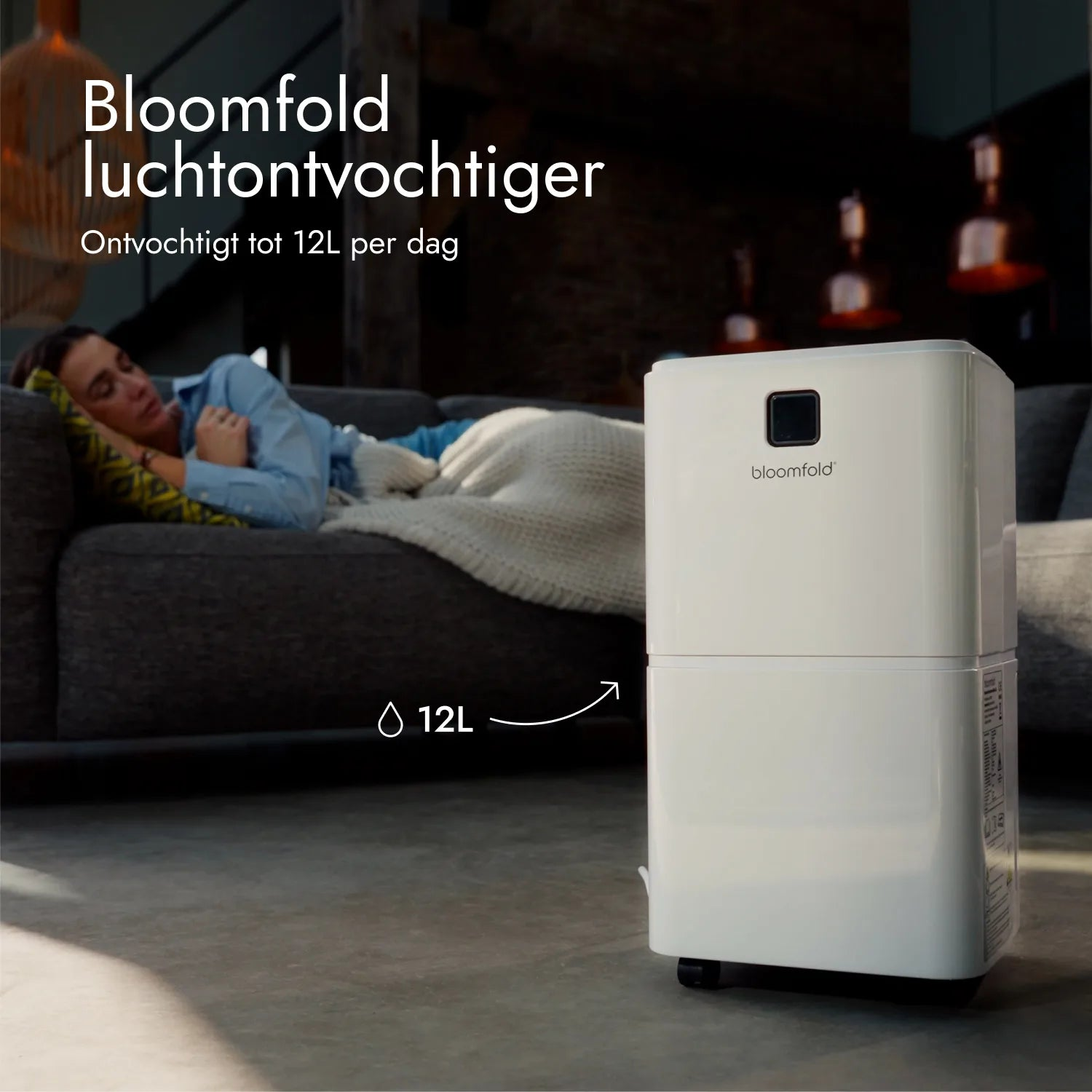 Ein weißer Bloomfold-Luftentfeuchter steht auf dem Boden, während eine Frau im Hintergrund auf dem Sofa schläft.
