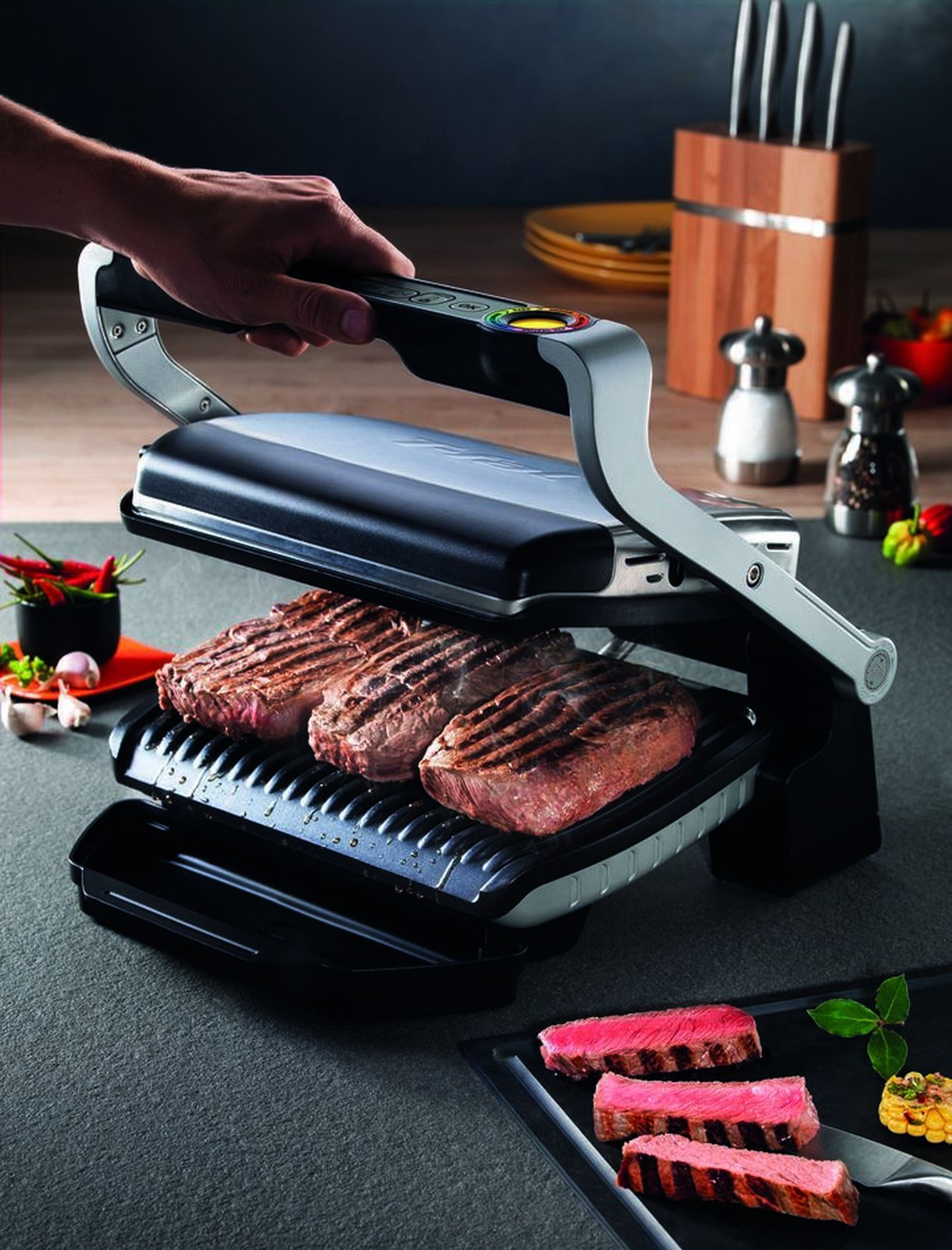 Ręka otwiera grill elektryczny z trzema stekami, a więcej pokrojonych steków znajduje się na blacie.