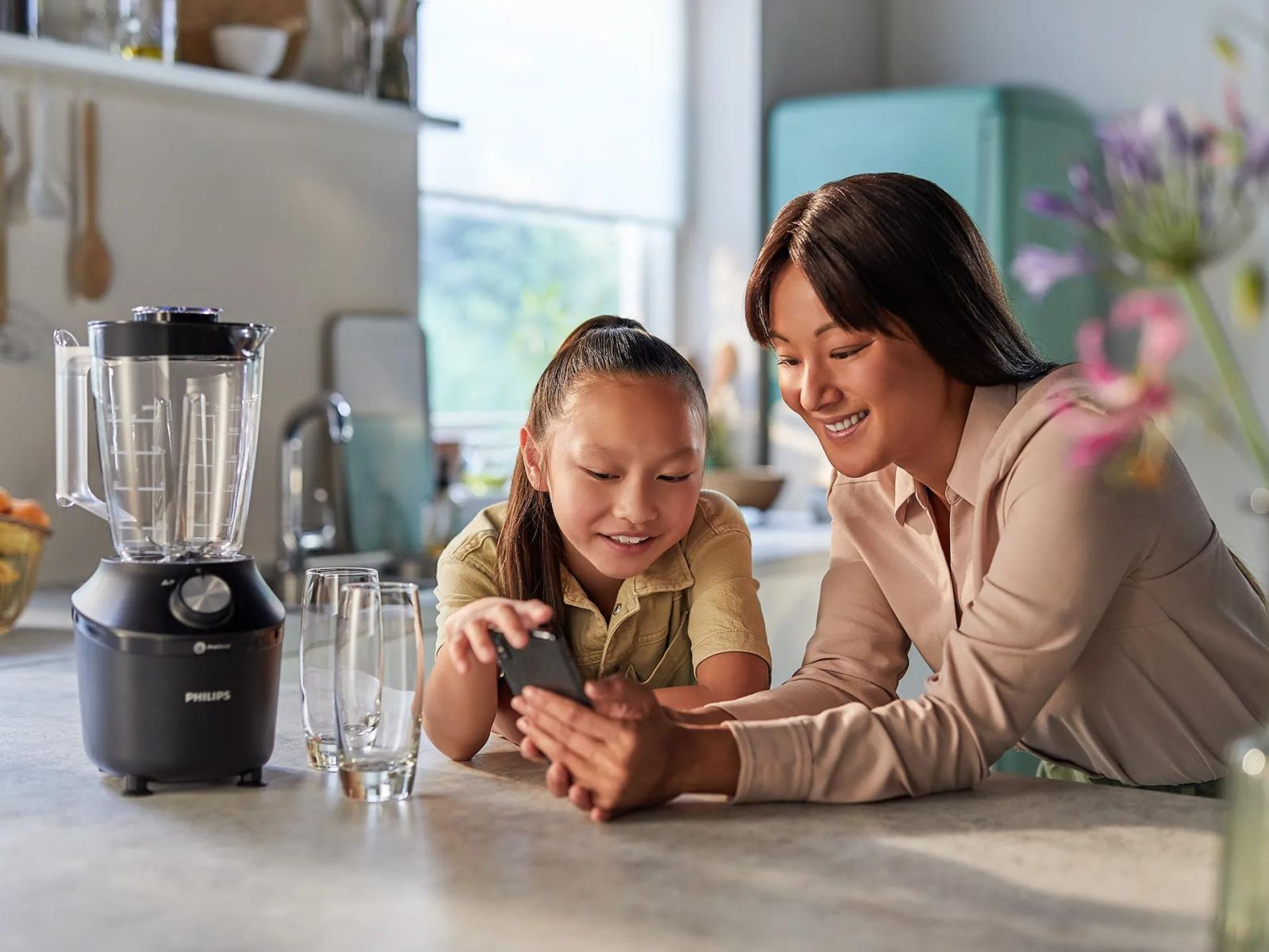 Una mujer y una niña sonrientes miran un teléfono inteligente en una cocina luminosa.