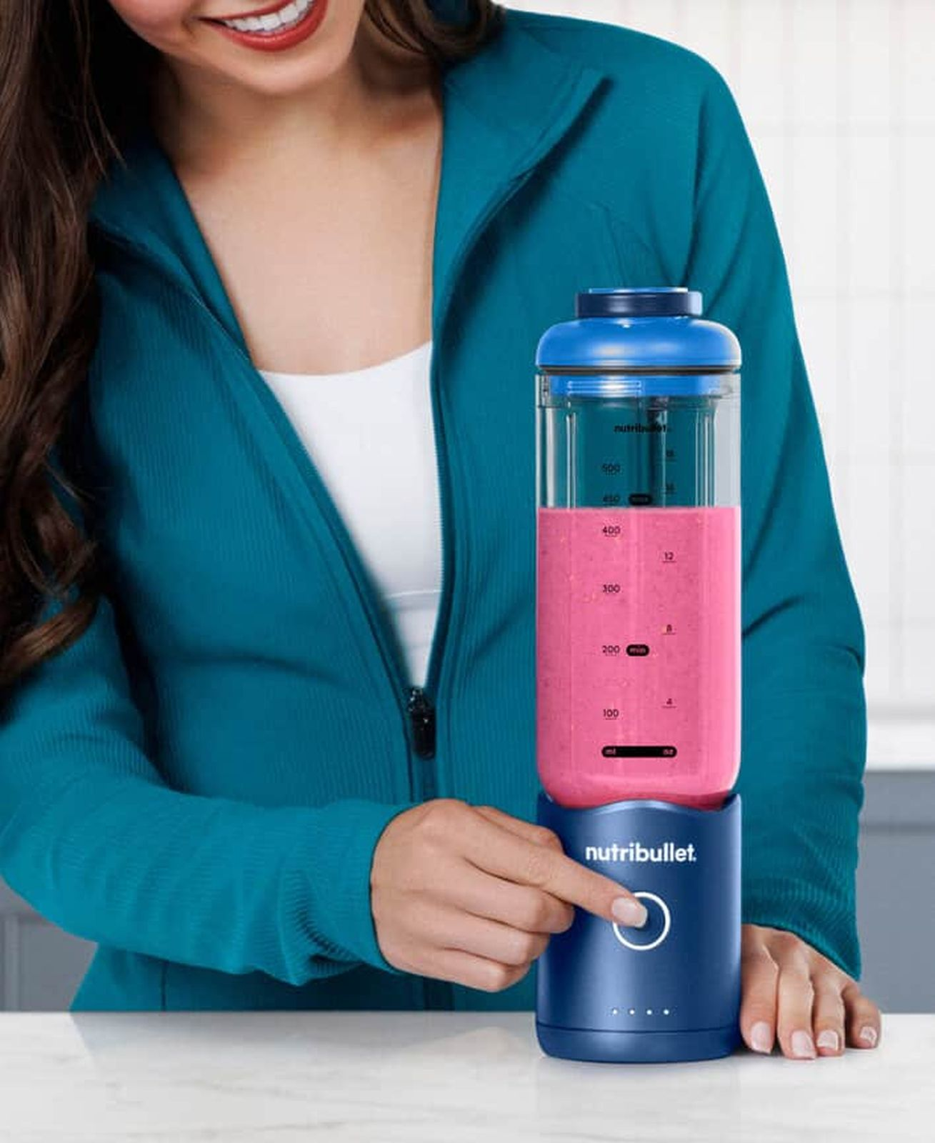Eine Frau in blauer Jacke drückt den Startknopf eines blauen Nutribullet-Mixers mit rosa Smoothie.