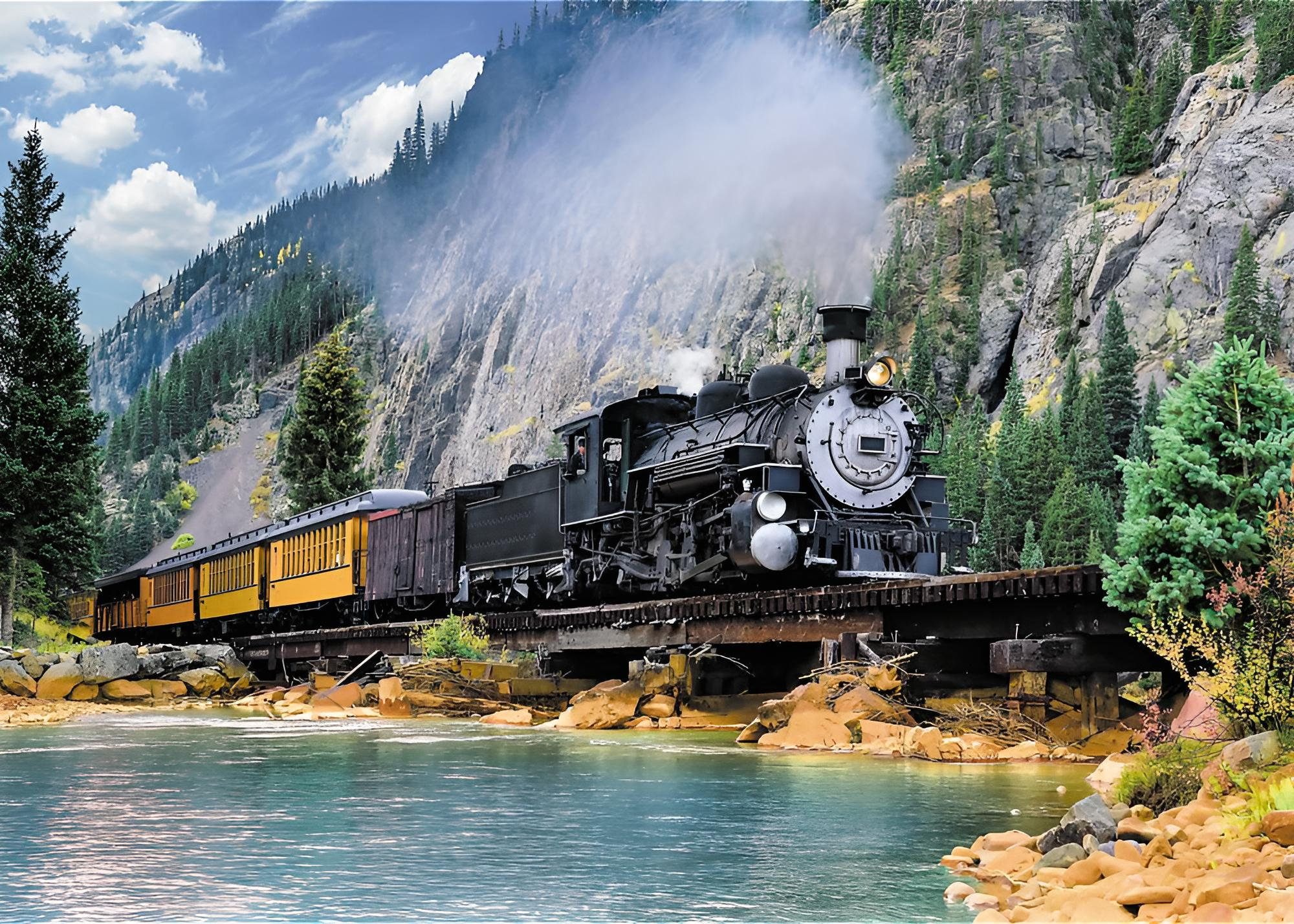 Un tren de vapor negro cruzando un puente de madera sobre un río, con montañas escarpadas y árboles de fondo.
