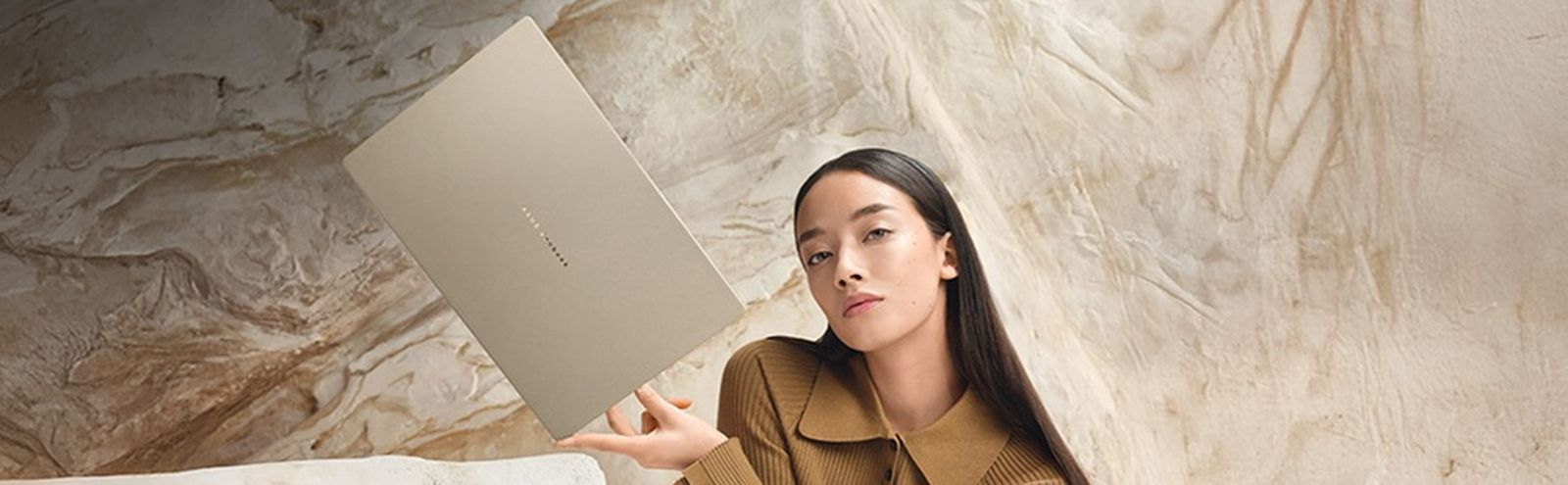 Eine Frau in einem braunen Pullover hält einen goldenen Laptop vor einer strukturierten Wand.