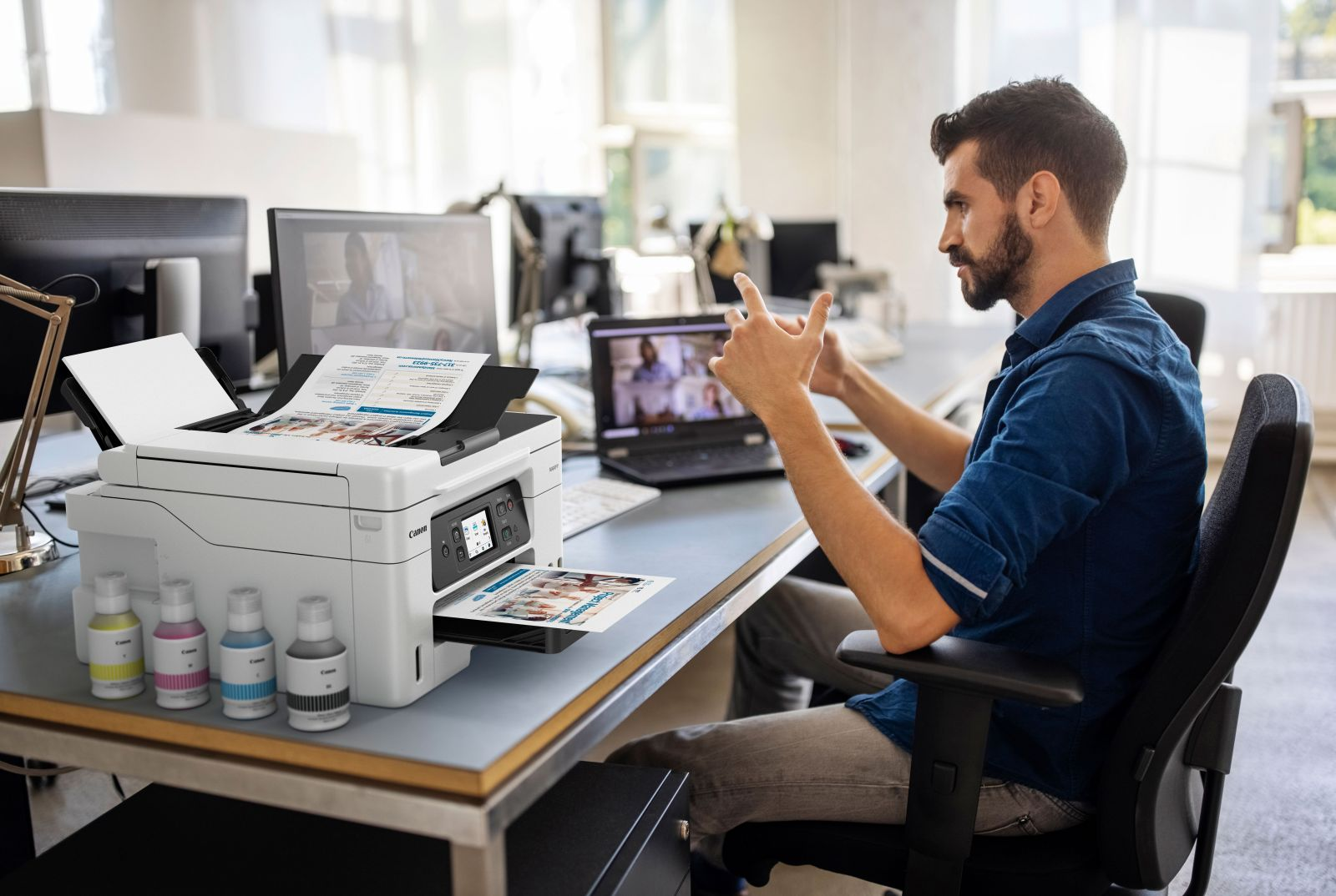 Un hombre con camisa azul está sentado en un escritorio de oficina, gesticulando ante una laptop y una impresora.