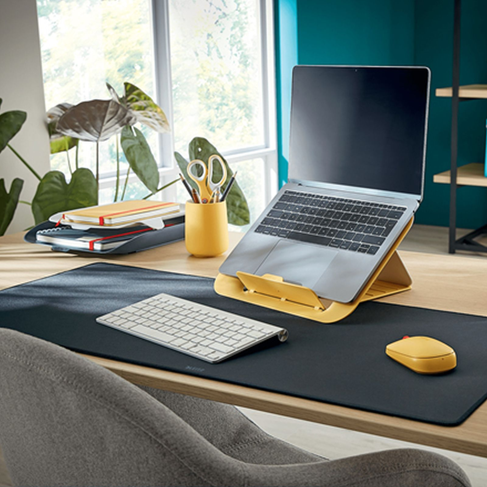Un escritorio con laptop en soporte amarillo, teclado blanco, ratón amarillo, cuadernos y plantas ante una ventana.