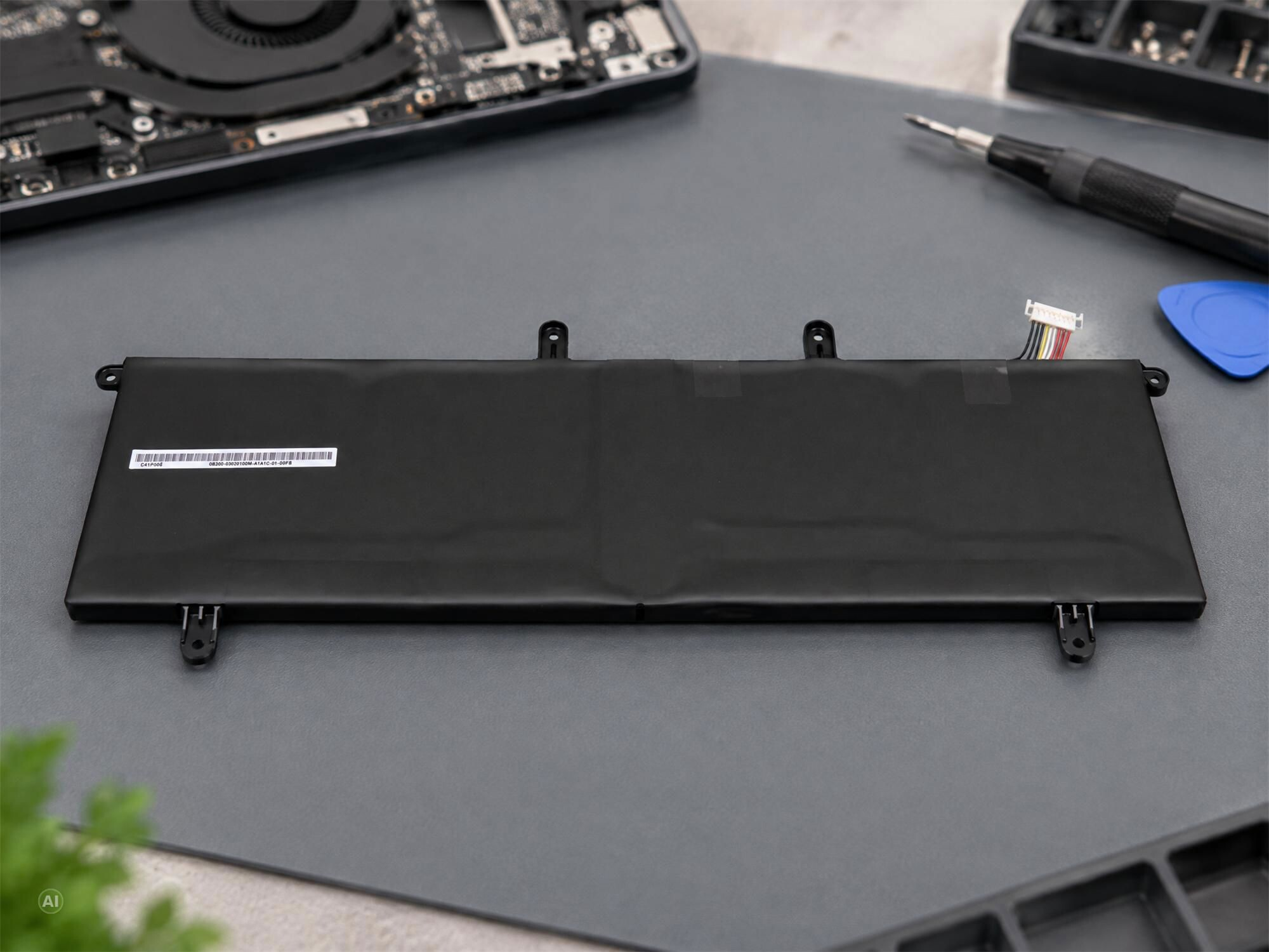 Ein schwarzer Laptop-Akku liegt auf einer grauen Werkbank, umgeben von Werkzeugen und Laptop-Teilen.