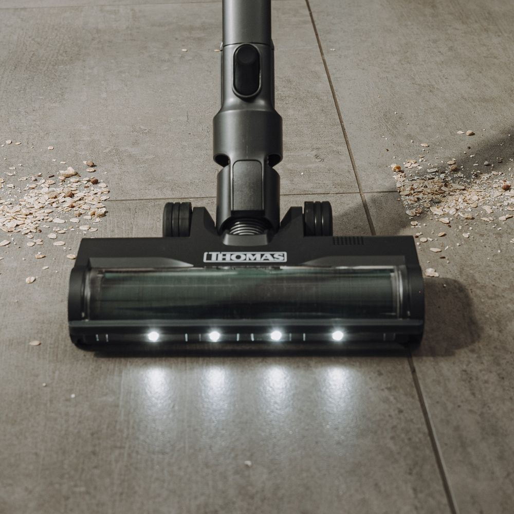 Un aspirapolvere nero con LED pulisce le briciole dalle piastrelle grigie.