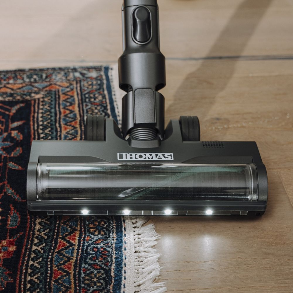 Una testa aspirapolvere Thomas nera con luci LED luminose su pavimento in legno e tappeto.