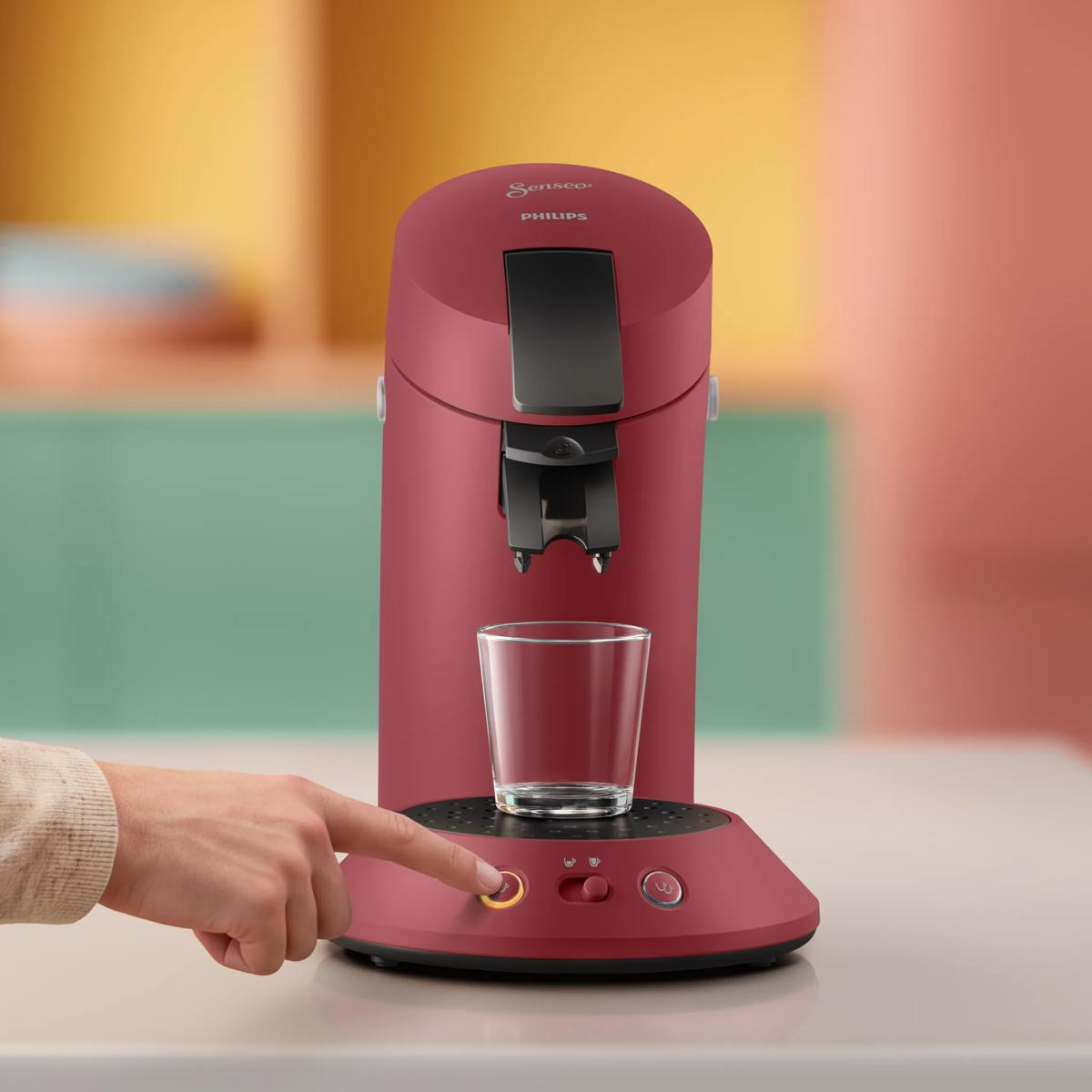 Una caffettiera rossa Philips Senseo con una tazza di vetro è su un tavolo, una mano preme il pulsante di avvio.