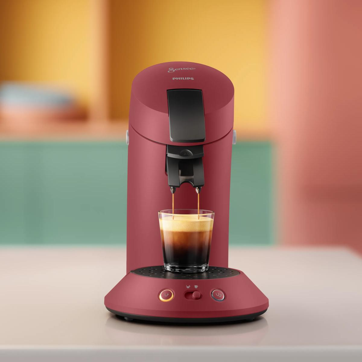 Una macchina da caffè Philips Senseo rossa eroga caffè in un bicchiere su una superficie chiara.