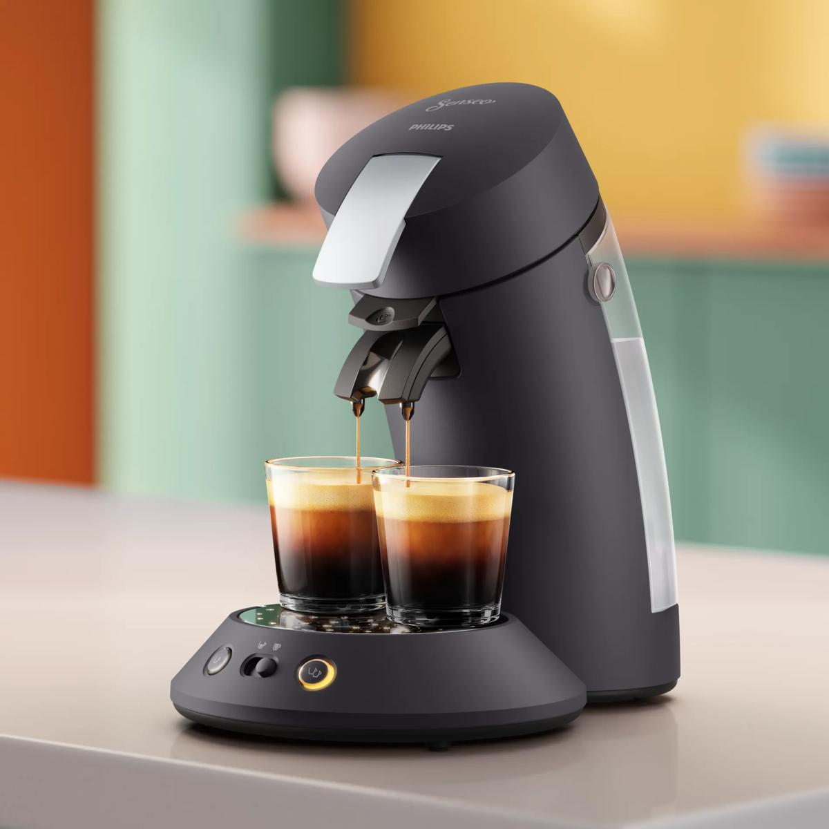 Cafetera Philips Senseo negra en un mostrador llenando dos vasos de café sobre un fondo pastel.
