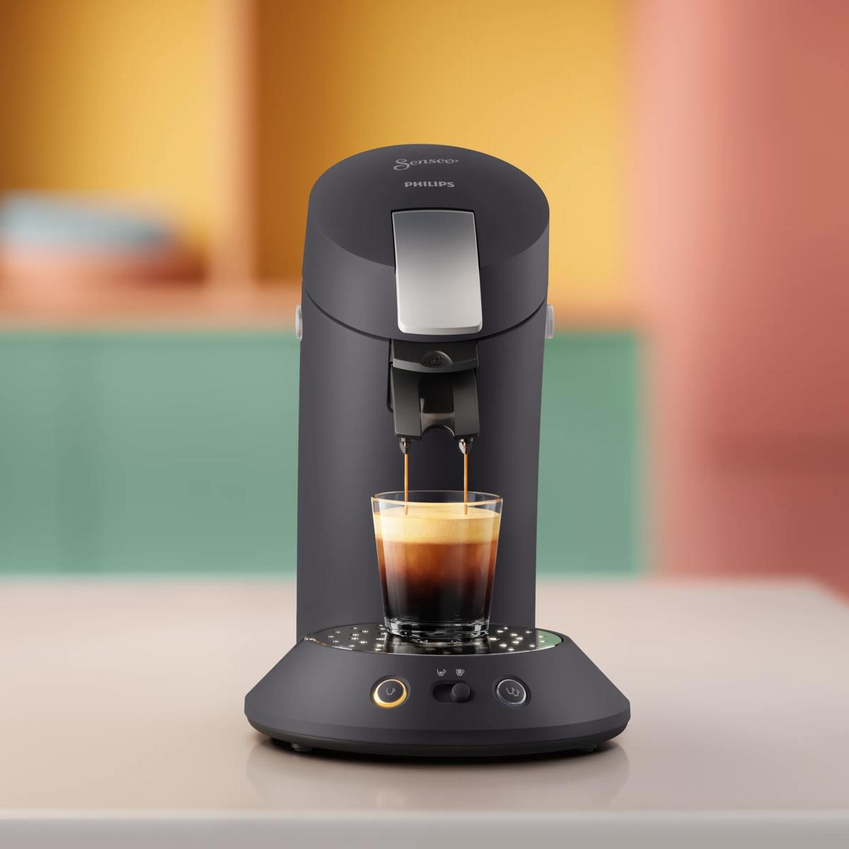 Una cafetera Philips Senseo negra prepara café en un vaso transparente sobre una encimera ante una pared colorida.