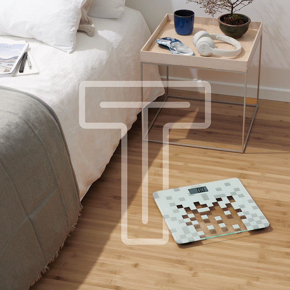 Un pèse-personne numérique blanc à motifs carrés repose sur le sol en bois d'une chambre.