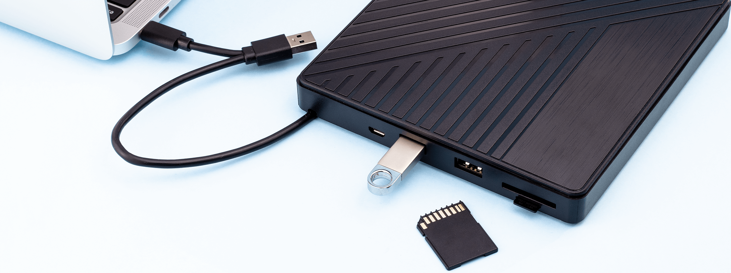Un hub esterno nero è collegato a un laptop e a una chiavetta USB, con una scheda SD vicina su sfondo azzurro.