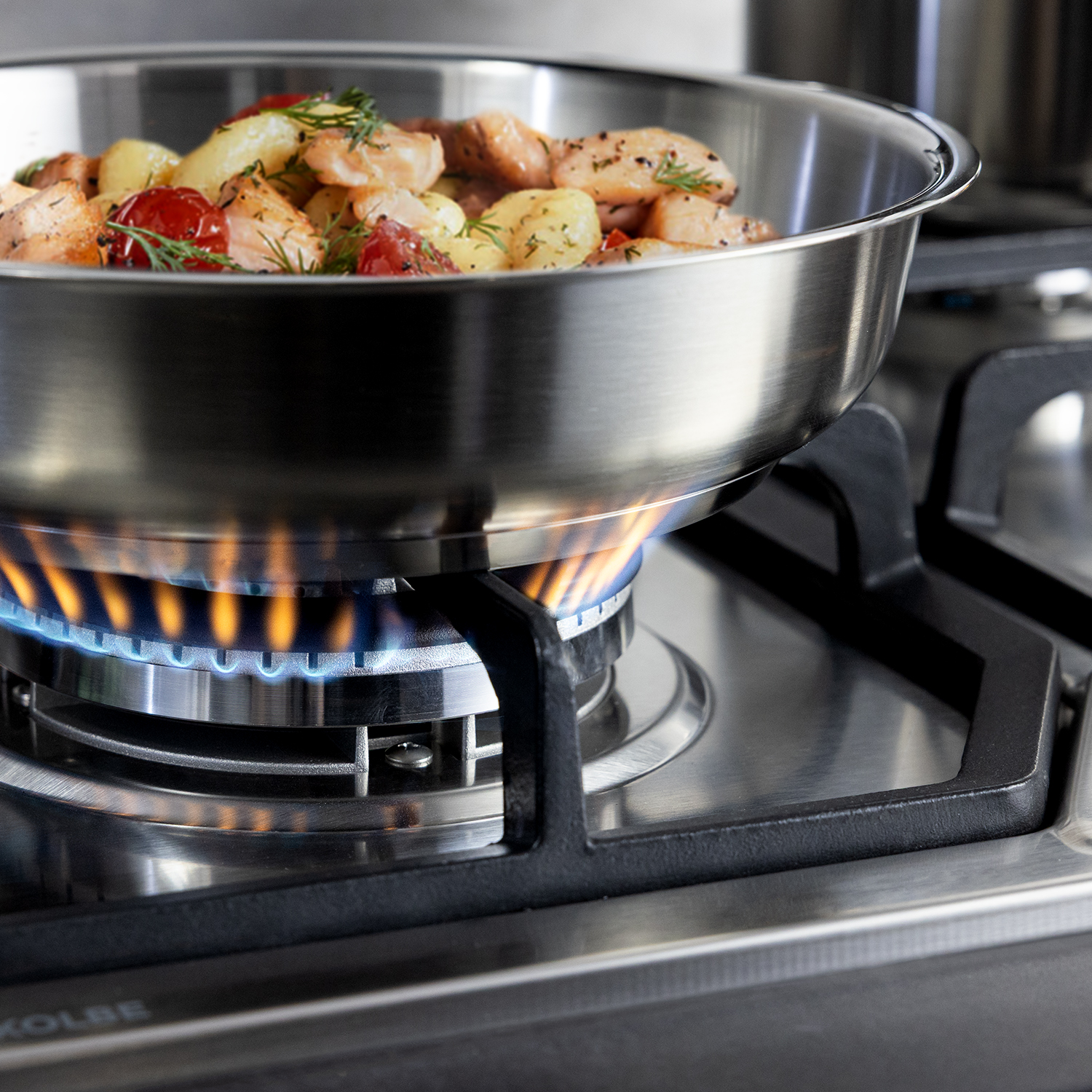 Eine Edelstahlpfanne mit gebratenem Lachs und Kartoffeln steht auf einer blauen Gasflamme.