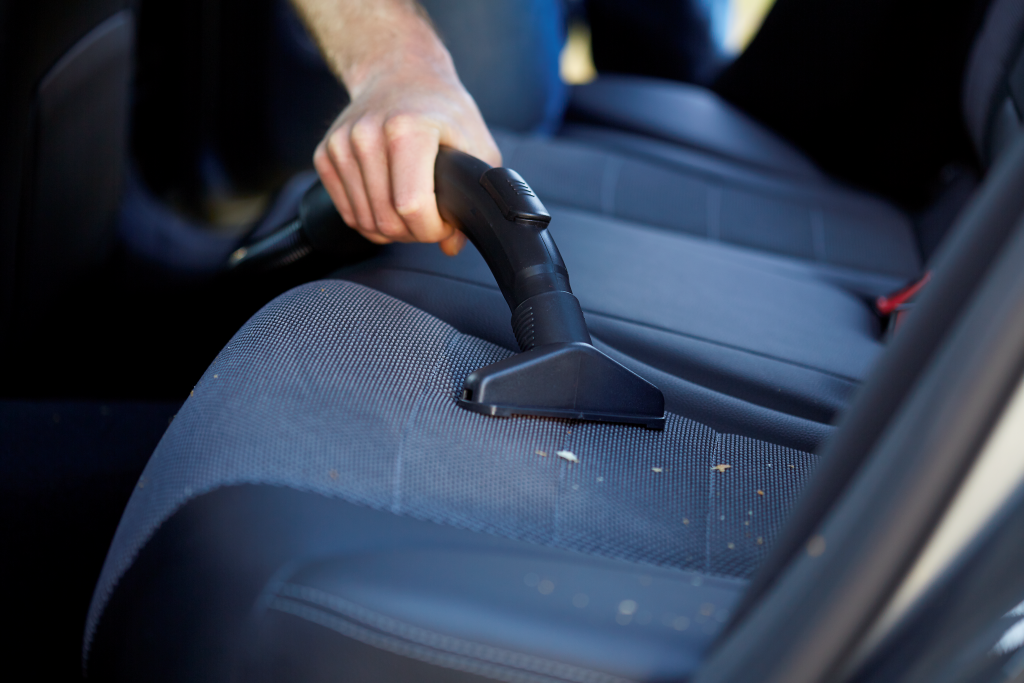 Une main aspire des miettes d'un siège de voiture sombre à l'aide d'un petit accessoire.