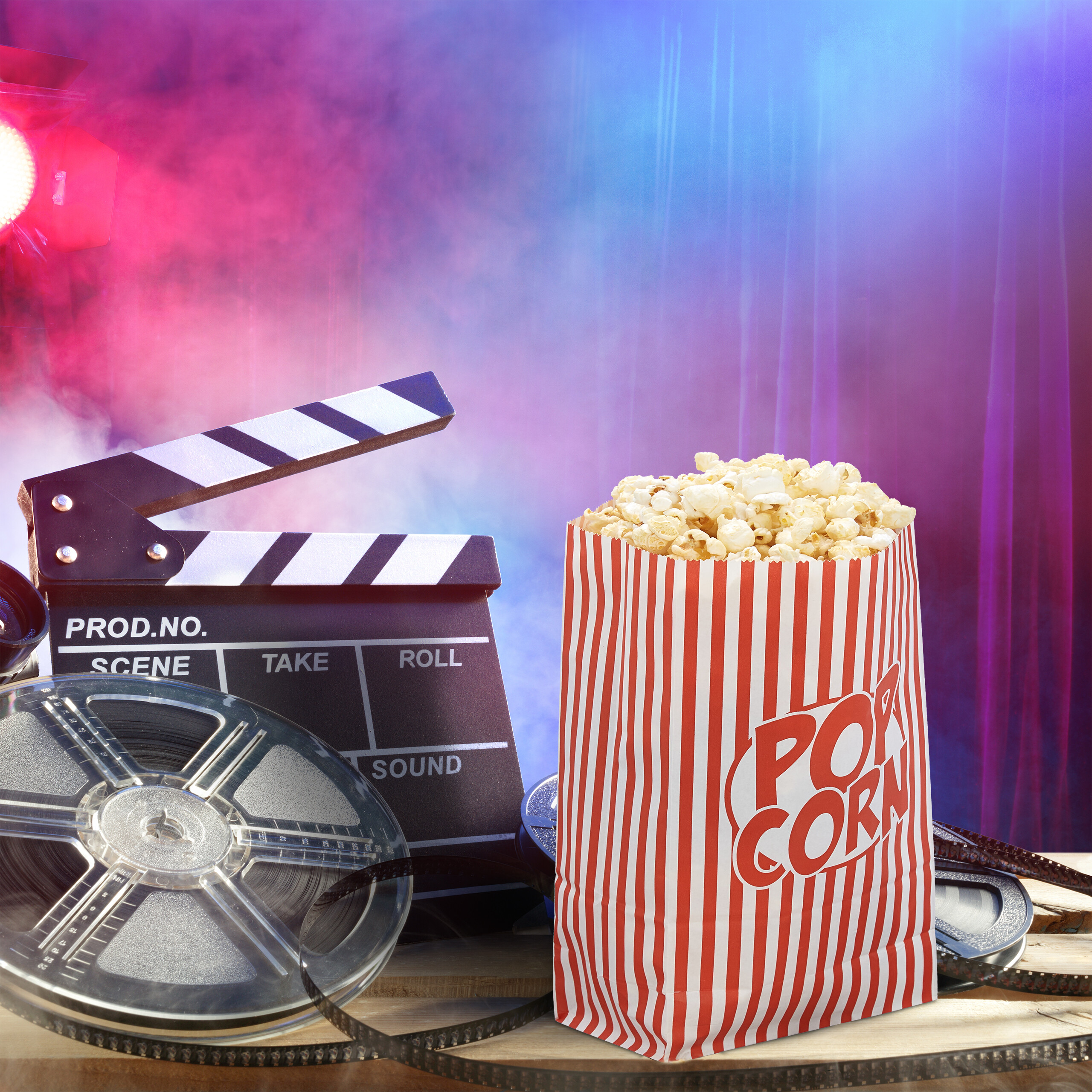 Popcorntüte, Filmklappe und Filmrolle auf Holztisch vor blau-rosa Hintergrund.
