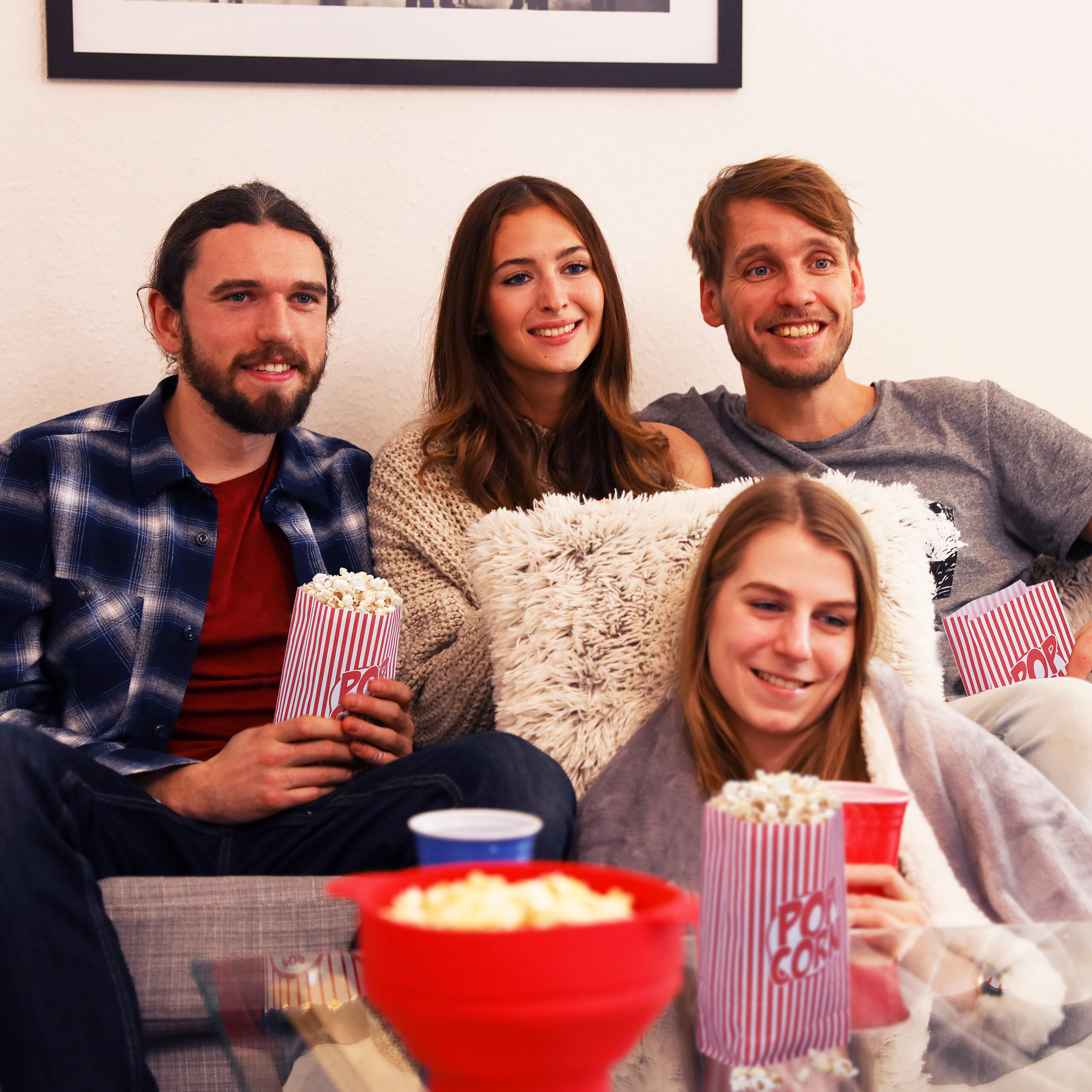 Vier junge Freunde sitzen zusammen auf einem Sofa, essen Popcorn und schauen vergnügt in eine Richtung.