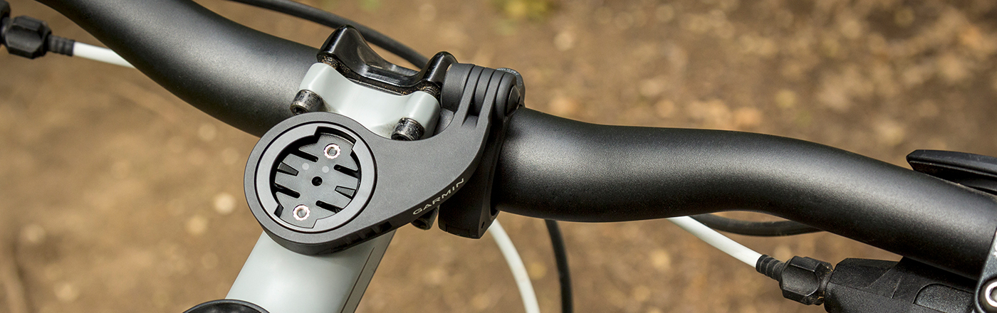Nahaufnahme eines Fahrradlenkers aus schwarzem Metall mit einer Garmin-Halterung am Vorbau vor unschärferem Boden.