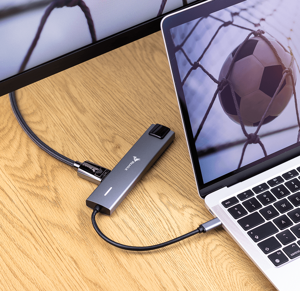 Ein grauer USB-C-Hub liegt auf einem Holztisch und ist mit einem Laptop und einem Monitor verbunden.
