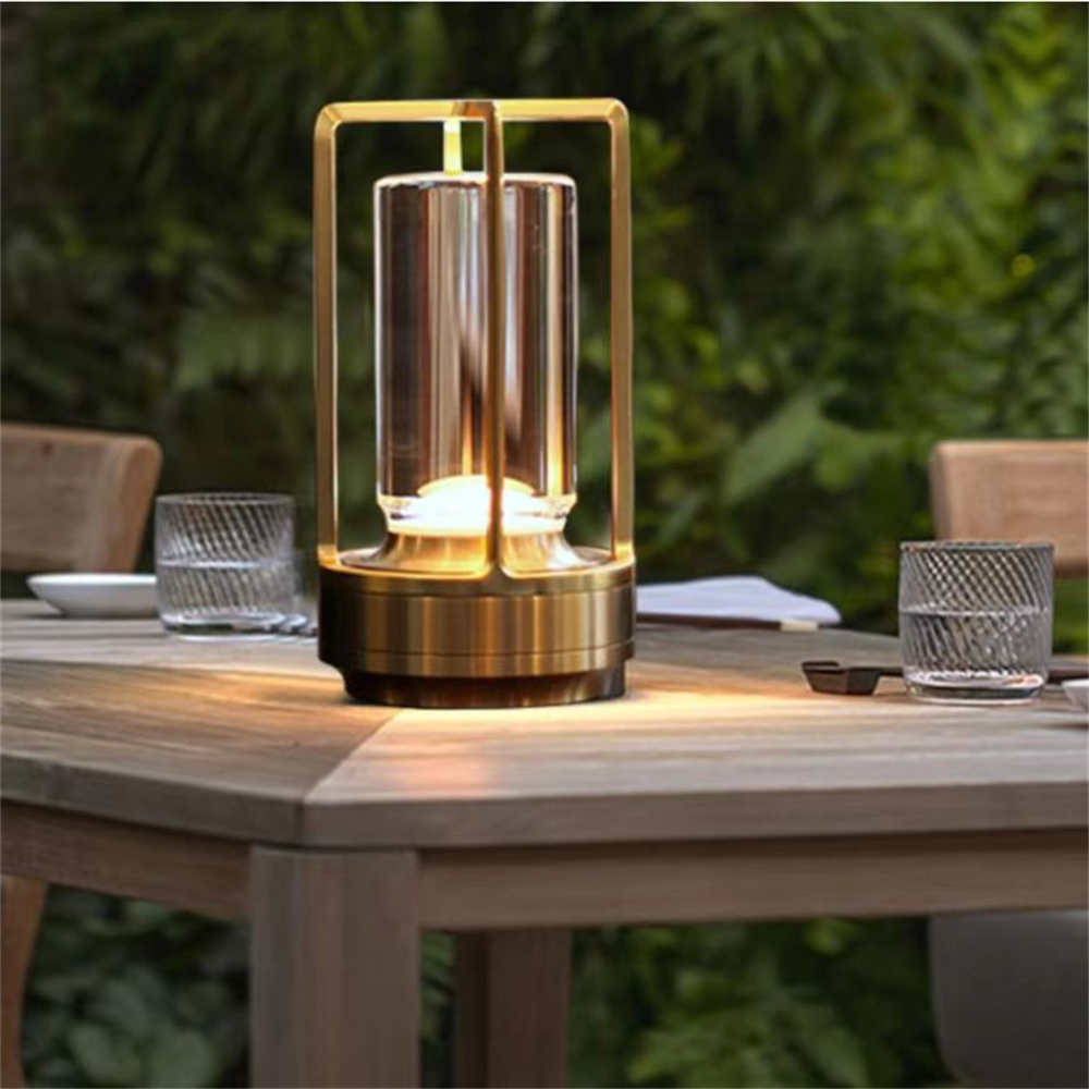 Een gouden tafellamp met glazen cilinder staat op een houten tafel buiten, omringd door groen.