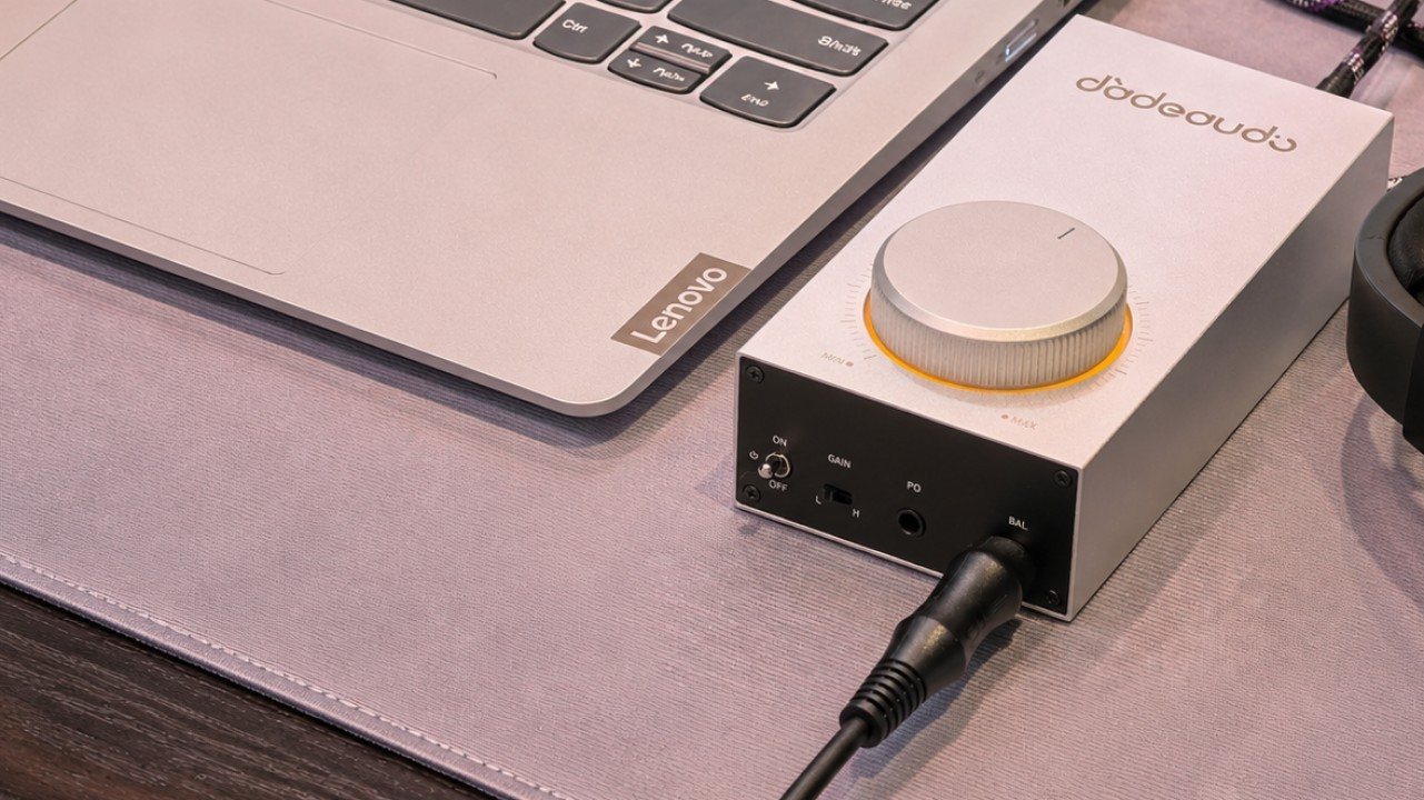 Srebrny wzmacniacz audio z dużym pokrętłem głośności stoi na biurku obok laptopa Lenovo i kabla.