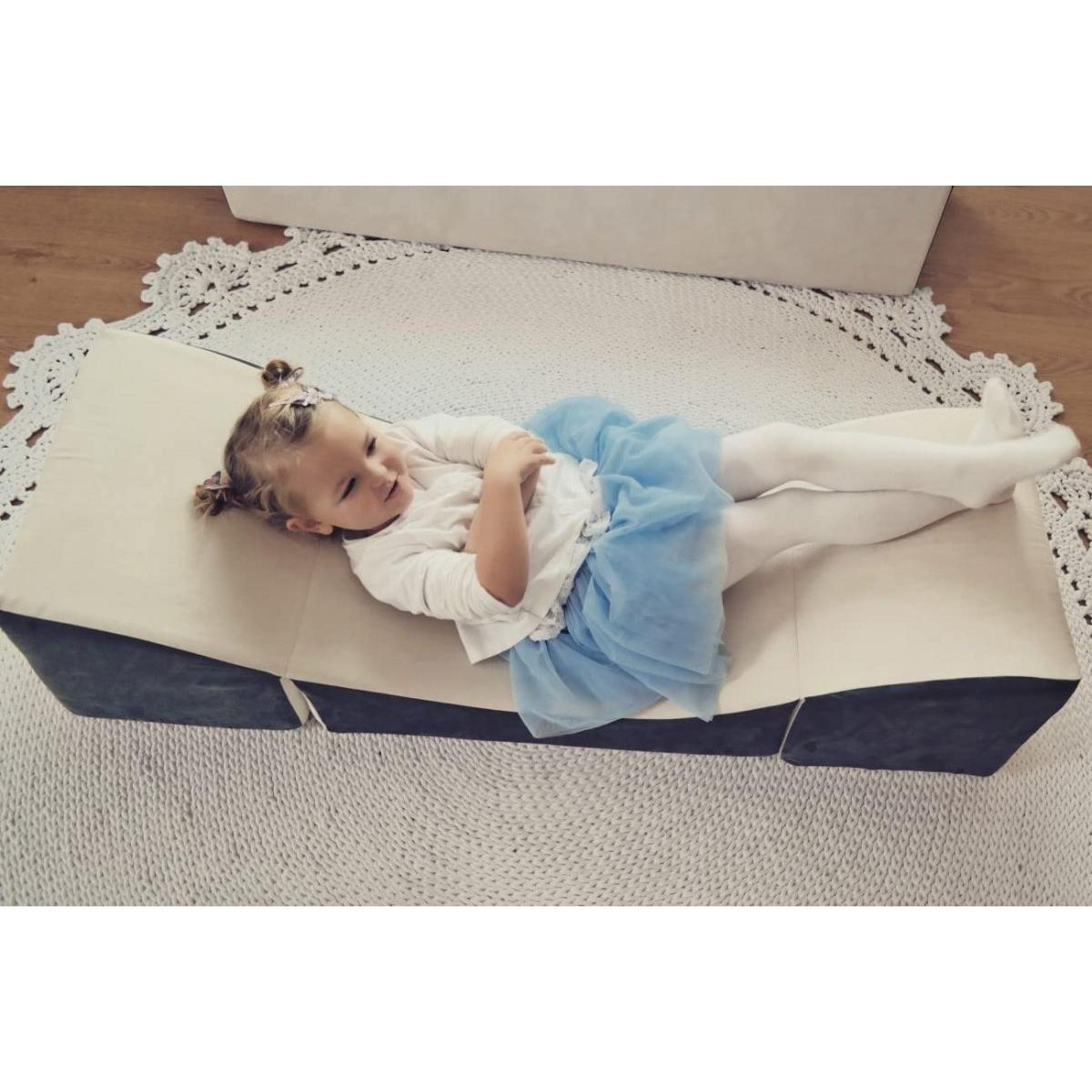 Kleines Mädchen in blauem Tutu liegt entspannt auf einem hellen Kissensofa über einem runden, weißen Teppich.