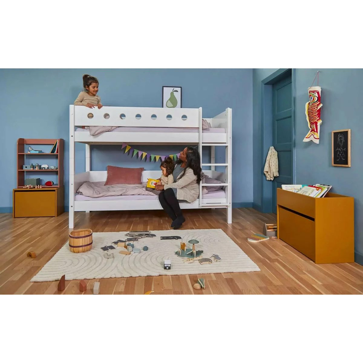 Eine Frau und zwei Kinder im hellblauen Kinderzimmer mit weißem Etagenbett, Holzboden und Spielzeug.