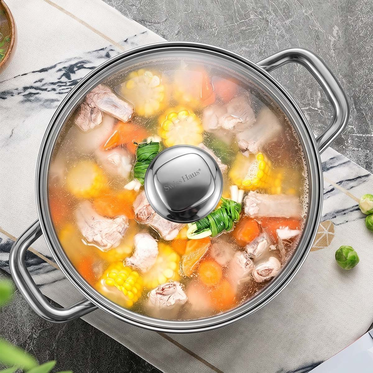 Ein großer edelstahltopf mit glasdeckel gefüllt mit suppe aus mais, karotten und fleisch auf einer tischdecke.