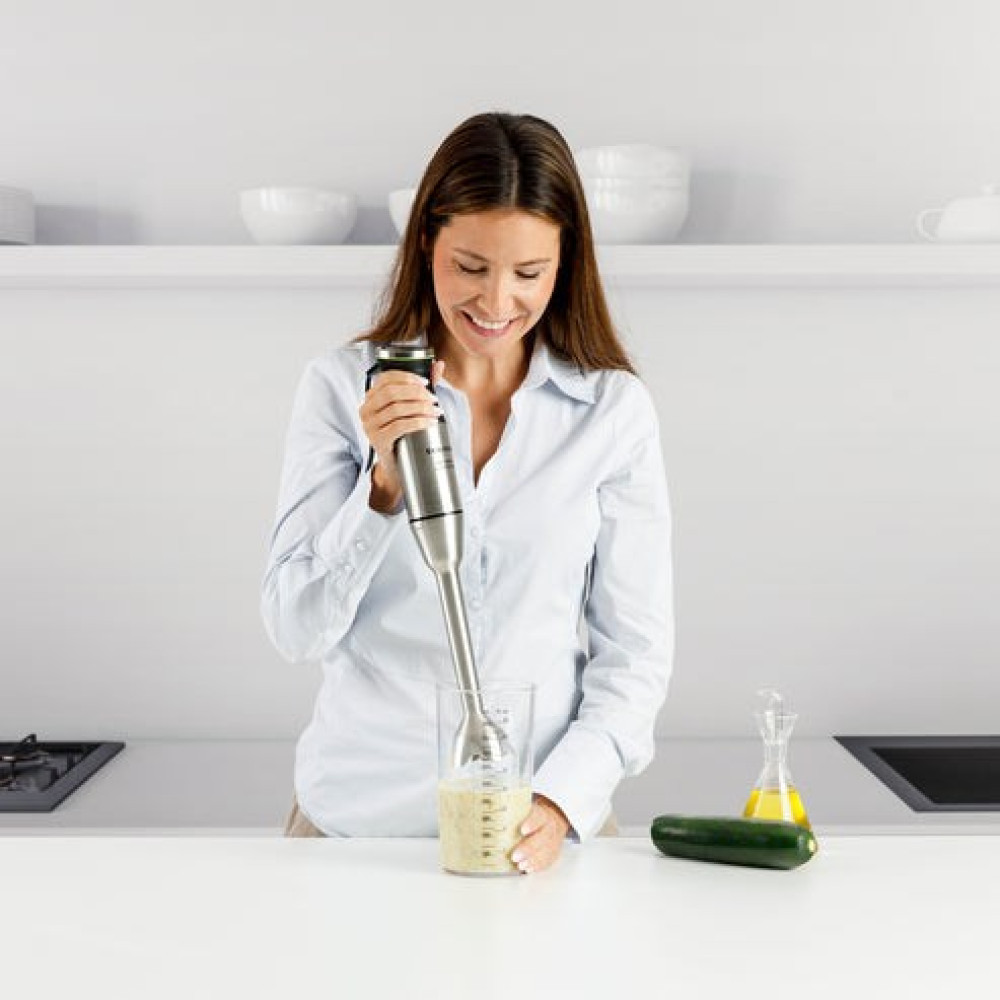 Una donna con camicia chiara usa un frullatore a immersione in una cucina con ripiani.