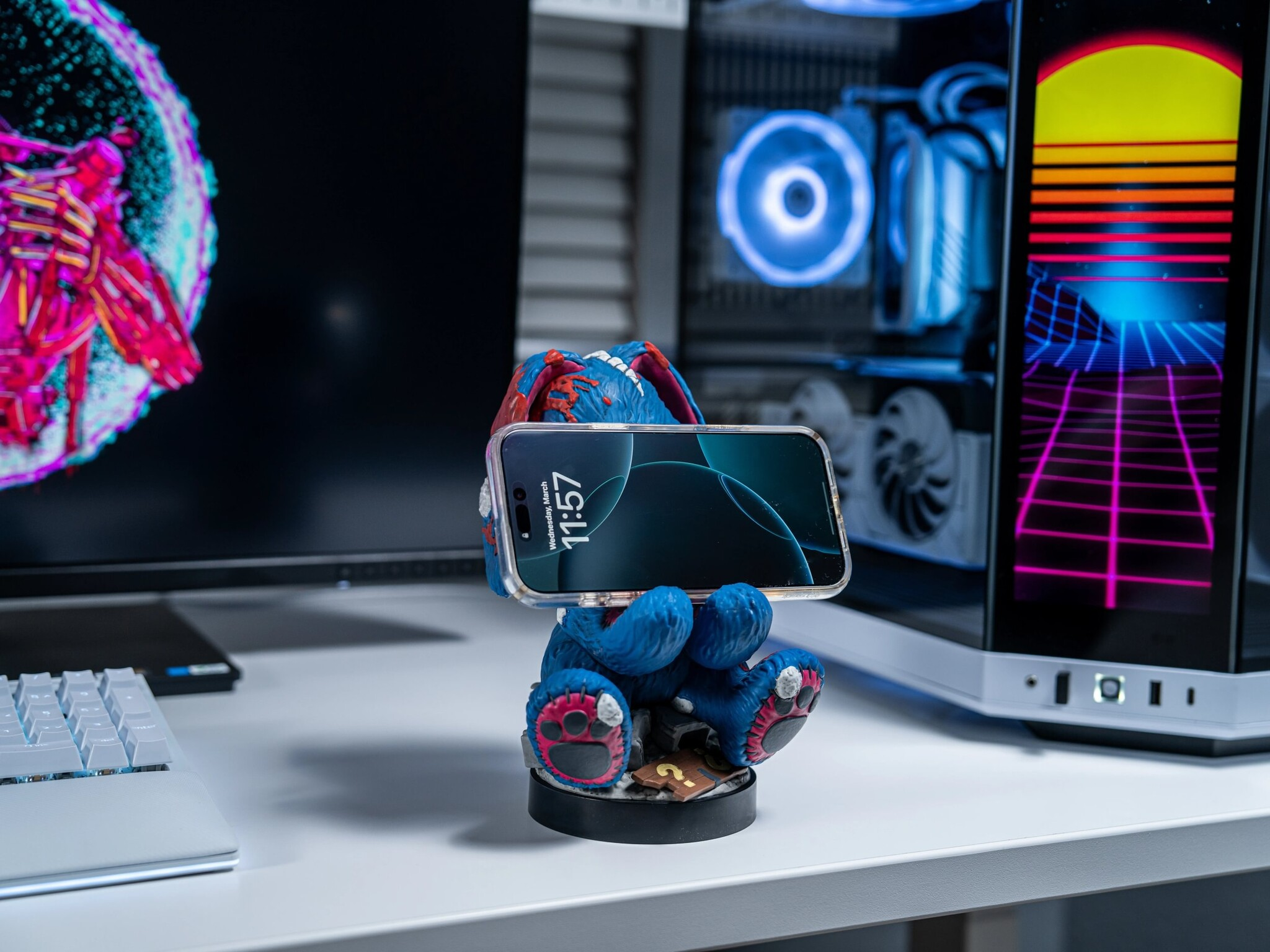Blaue Monsterfigur hält ein Smartphone auf einem weißen Schreibtisch mit Monitoren und einem PC im Hintergrund.