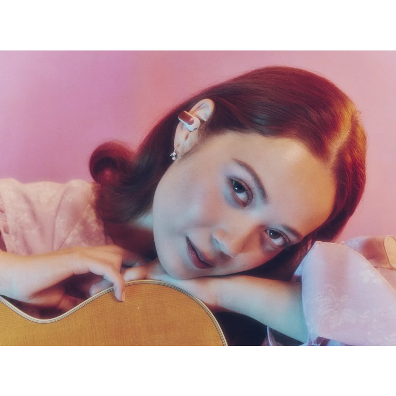 Eine Frau mit rotem Haar und Ohrstöpseln lehnt sich vor einem pinken Hintergrund an eine Gitarre.