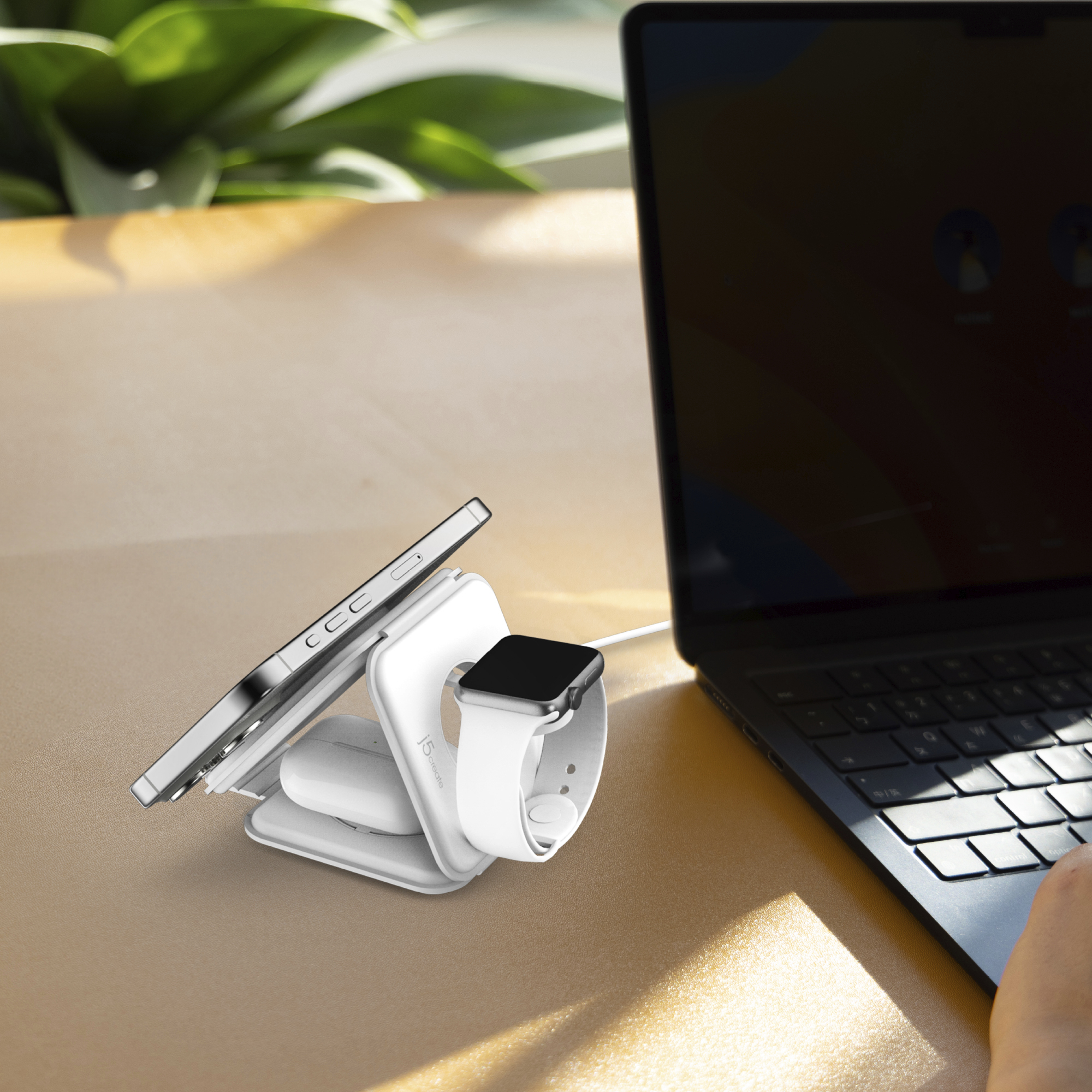 Eine weiße 3-in-1-Ladestation hält Smartphone, Ohrhörer und Smartwatch neben einem Laptop auf einem Holztisch.