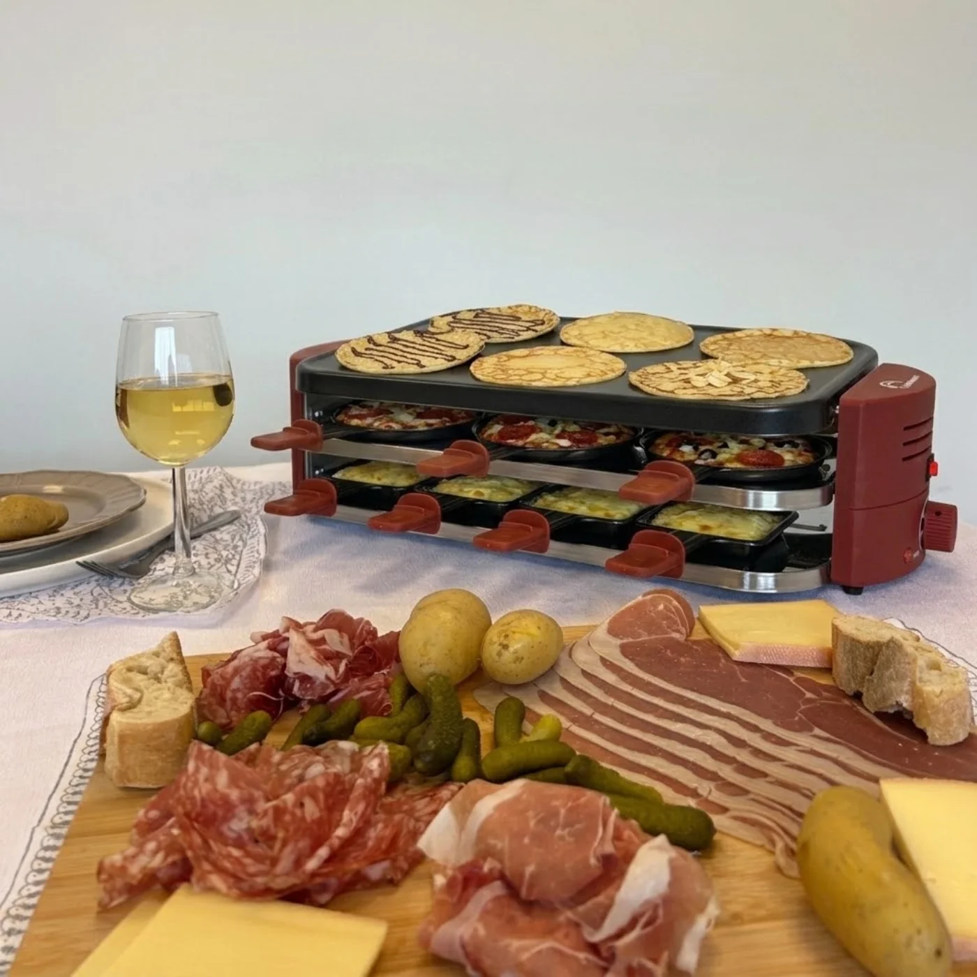 Ein Raclette-Grill mit Pfannkuchen und Mini-Pizzen, begleitet von Kartoffeln, Aufschnitt, Käse, Gurken und Wein.