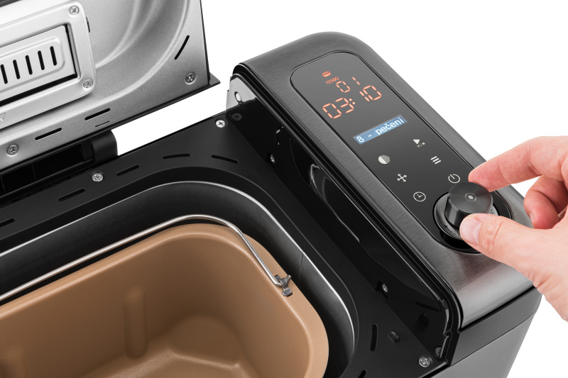 Eine Hand bedient den Drehknopf auf dem digitalen Bedienfeld eines offenen Brotbackautomaten mit brauner Backform.