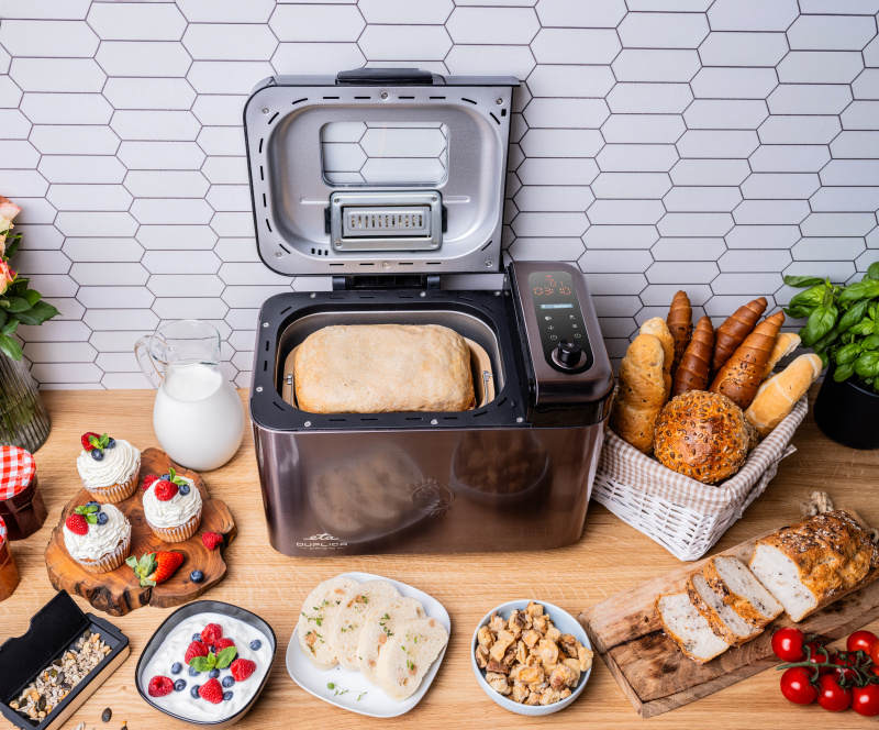 Brotbackautomat auf Holztheke mit Brot, Cupcakes, Milchkrug und Gebäck.