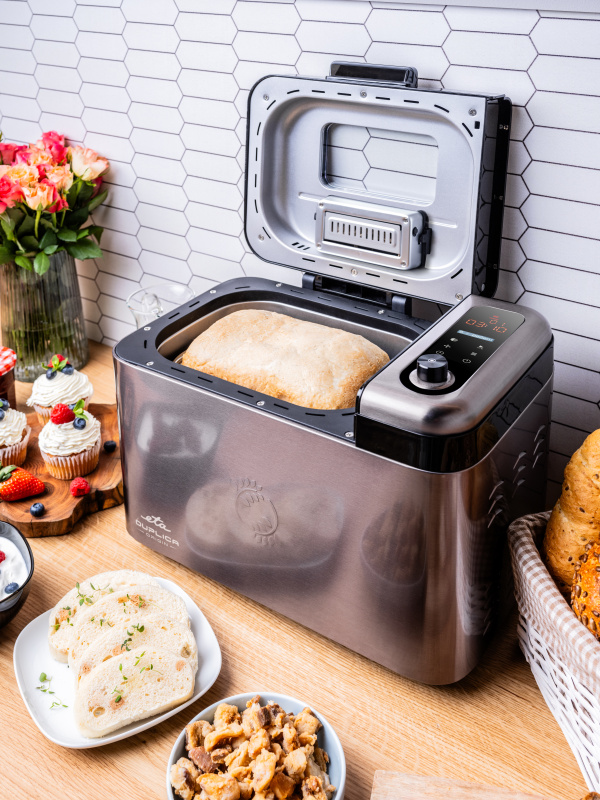 Ein silberner Brotbackautomat auf einer Holzarbeitsplatte, daneben Brot, Cupcakes und Blumen.