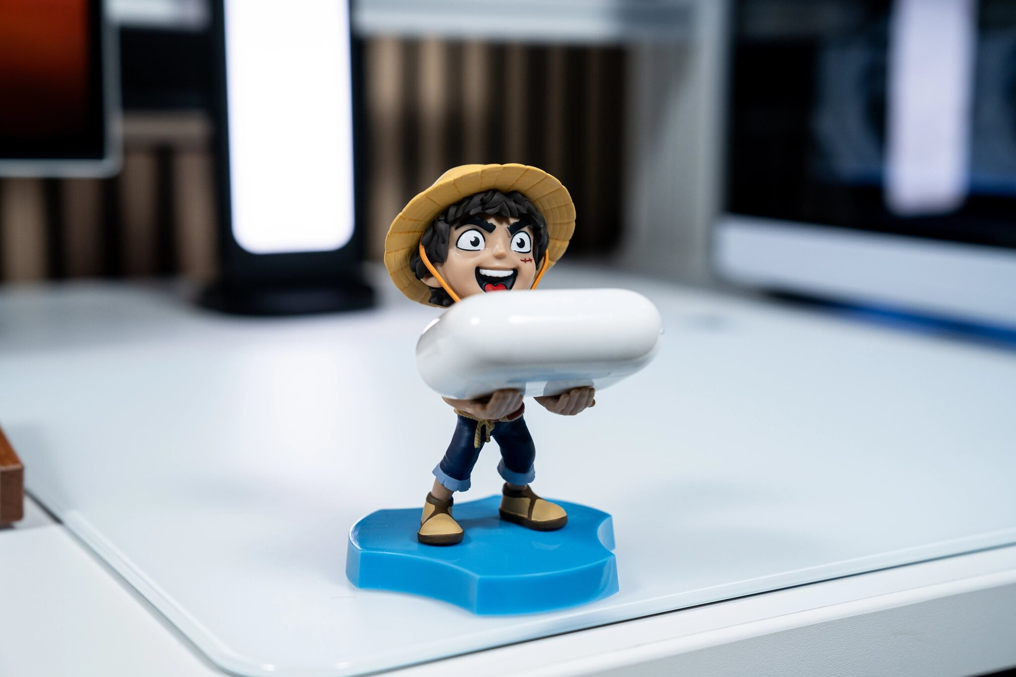 Una pequeña figura de Luffy con sombrero de paja que sostiene un estuche de auriculares blanco sobre una base azul en un escritorio.