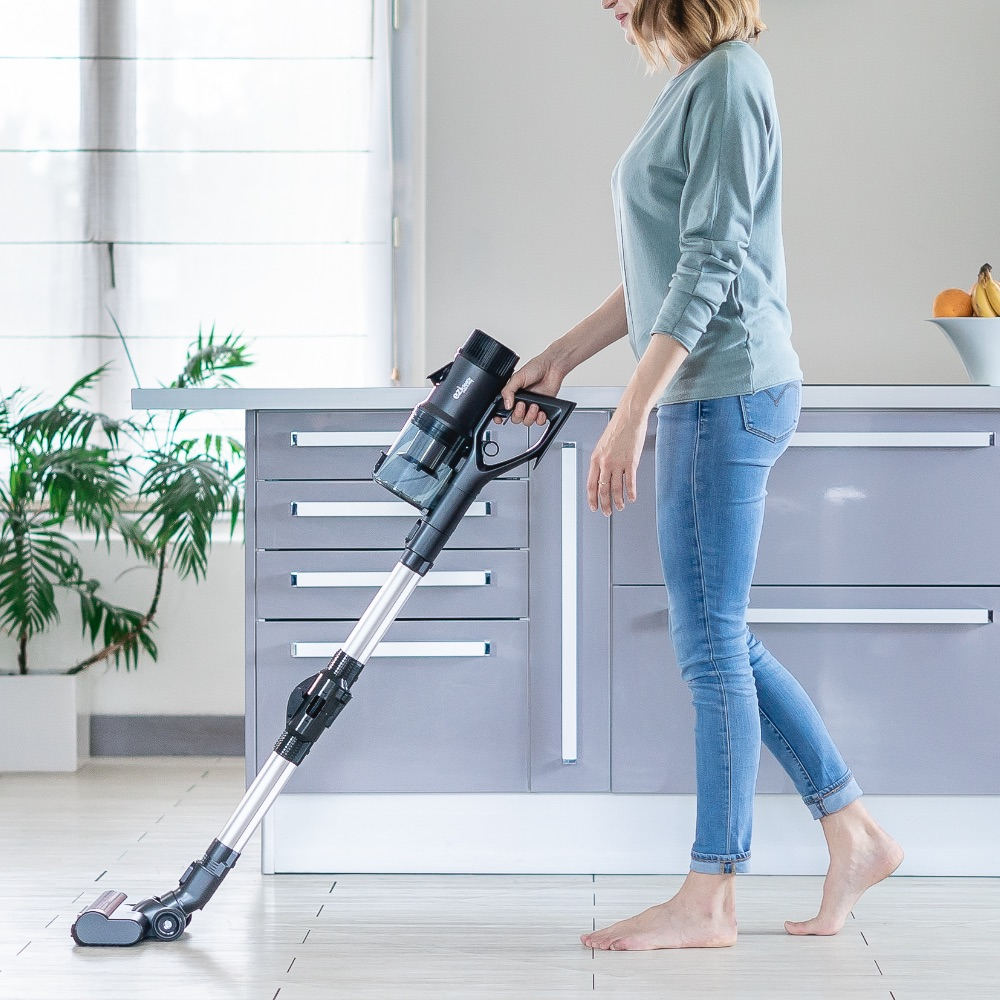 Eine Frau in Jeans und grünem Pullover saugt den Boden mit einem kabellosen Staubsauger in einer Küche.
