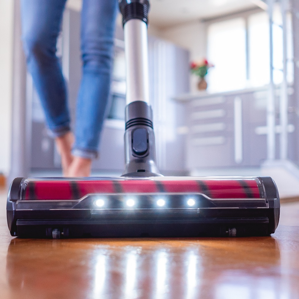 Ein Staubsauger mit leuchtenden LED-Lichtern reinigt einen Holzfußboden, im Hintergrund steht eine Person in Jeans.