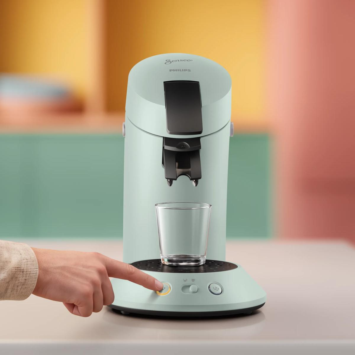 Una mano preme il pulsante di una macchina da caffè Philips Senseo verde menta con un bicchiere vuoto.