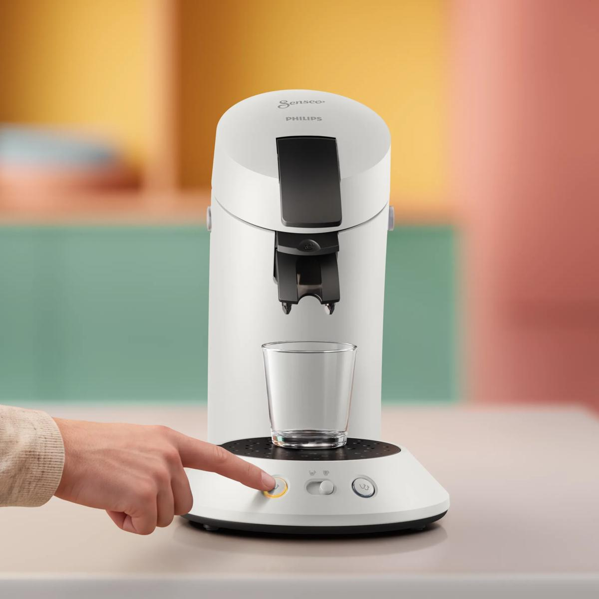 Una mano preme il pulsante di accensione di una macchina da caffè Philips Senseo bianca con una tazza.