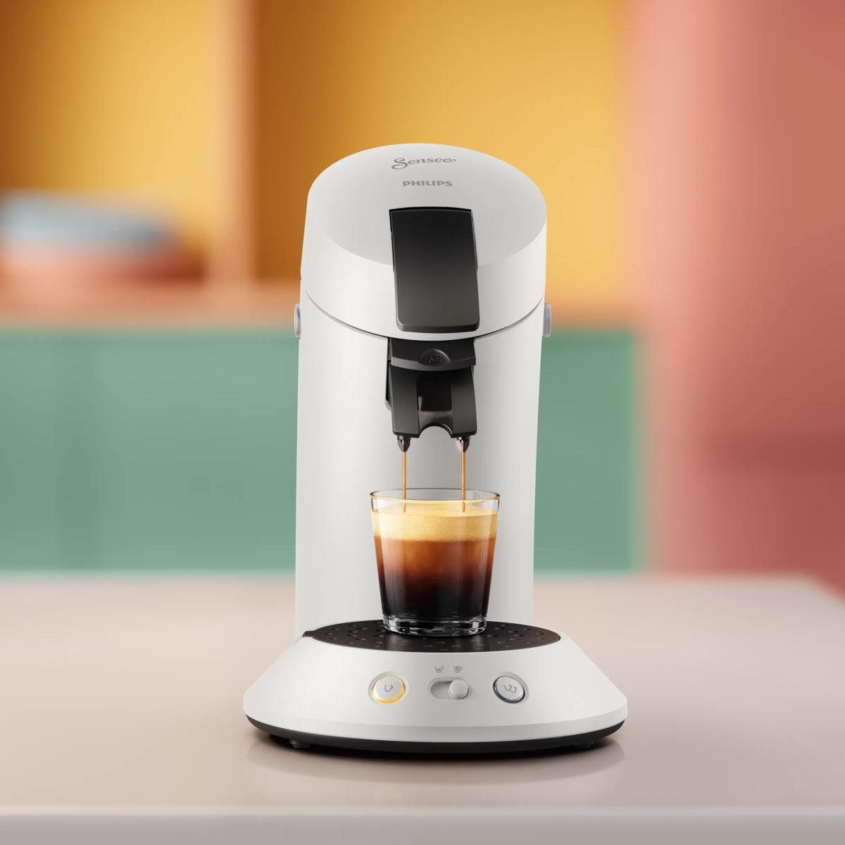Una macchina da caffè Philips Senseo bianca eroga caffè in un bicchiere su un bancone con sfondo pastello colorato.