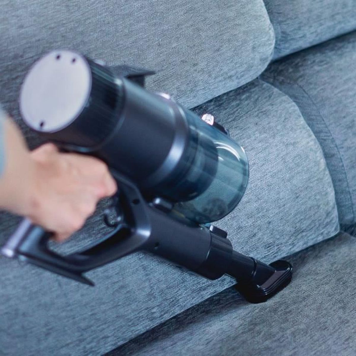 Eine Hand benutzt einen kabellosen Staubsauger, um ein graues Sofa zu reinigen.