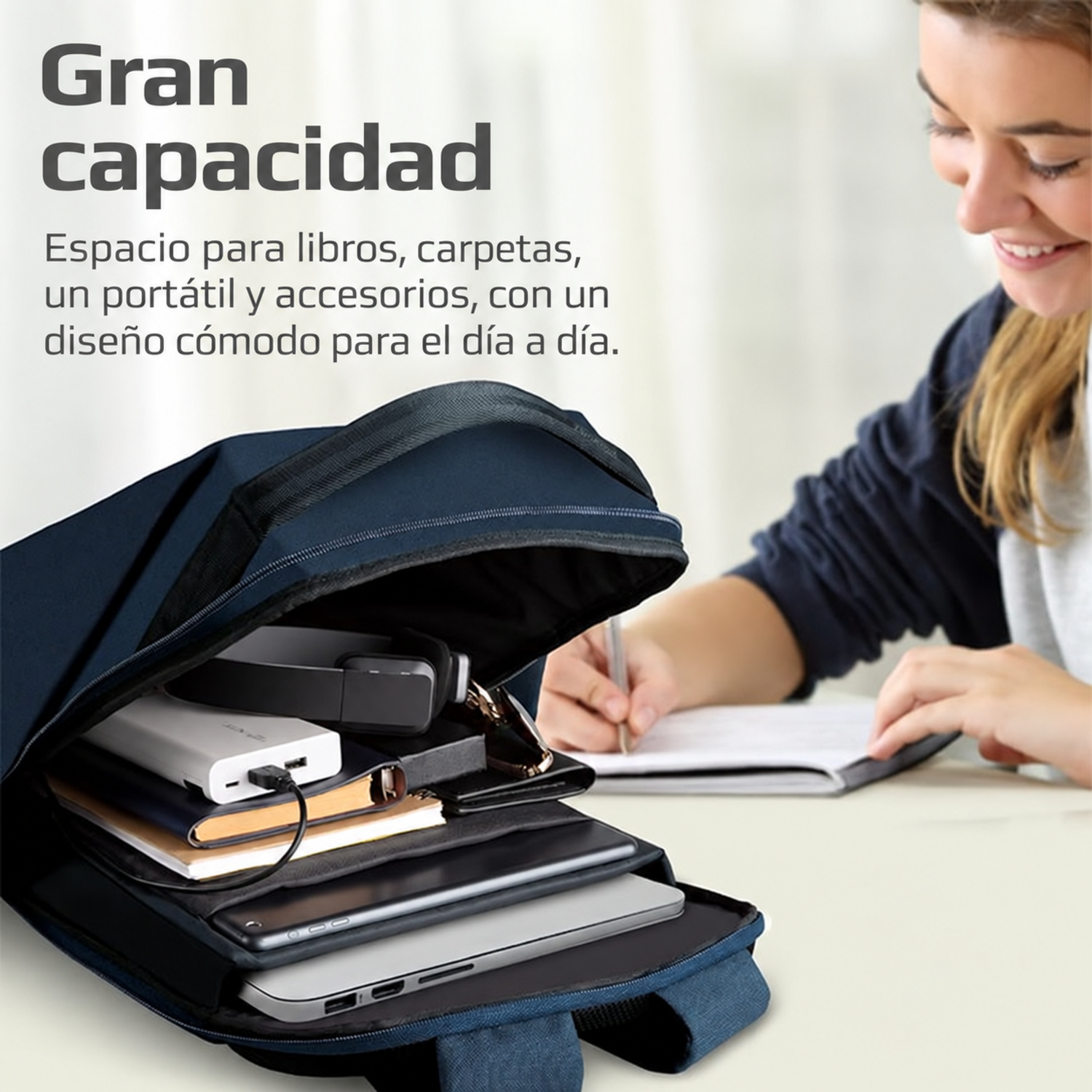 Una mochila azul abierta en la mesa con portátil, tablet y accesorios, y una mujer escribiendo al lado.