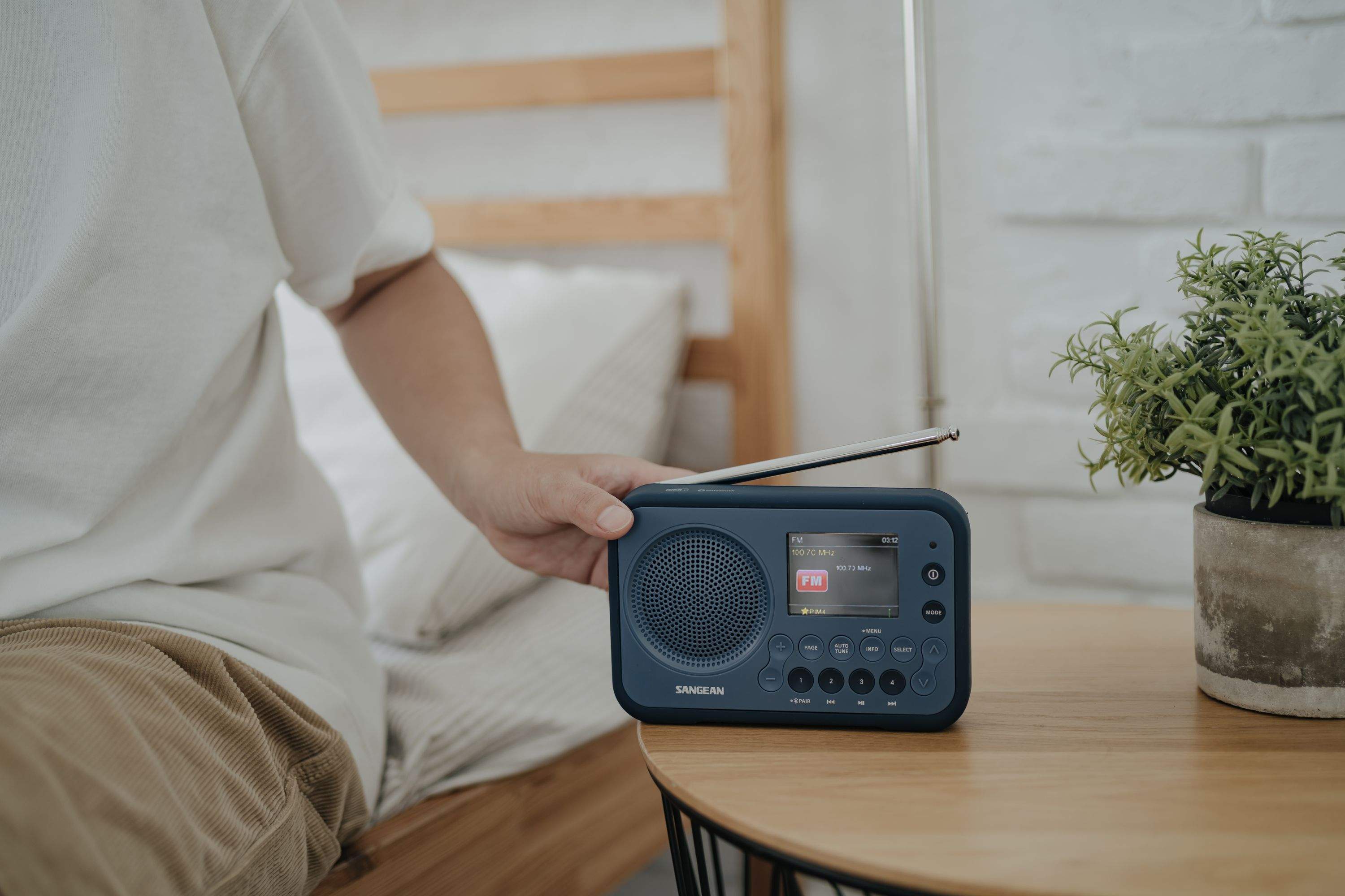 Eine Person sitzt auf dem Bett und berührt ein blaues Sangean-Radio auf einem Holztisch mit einer Pflanze.