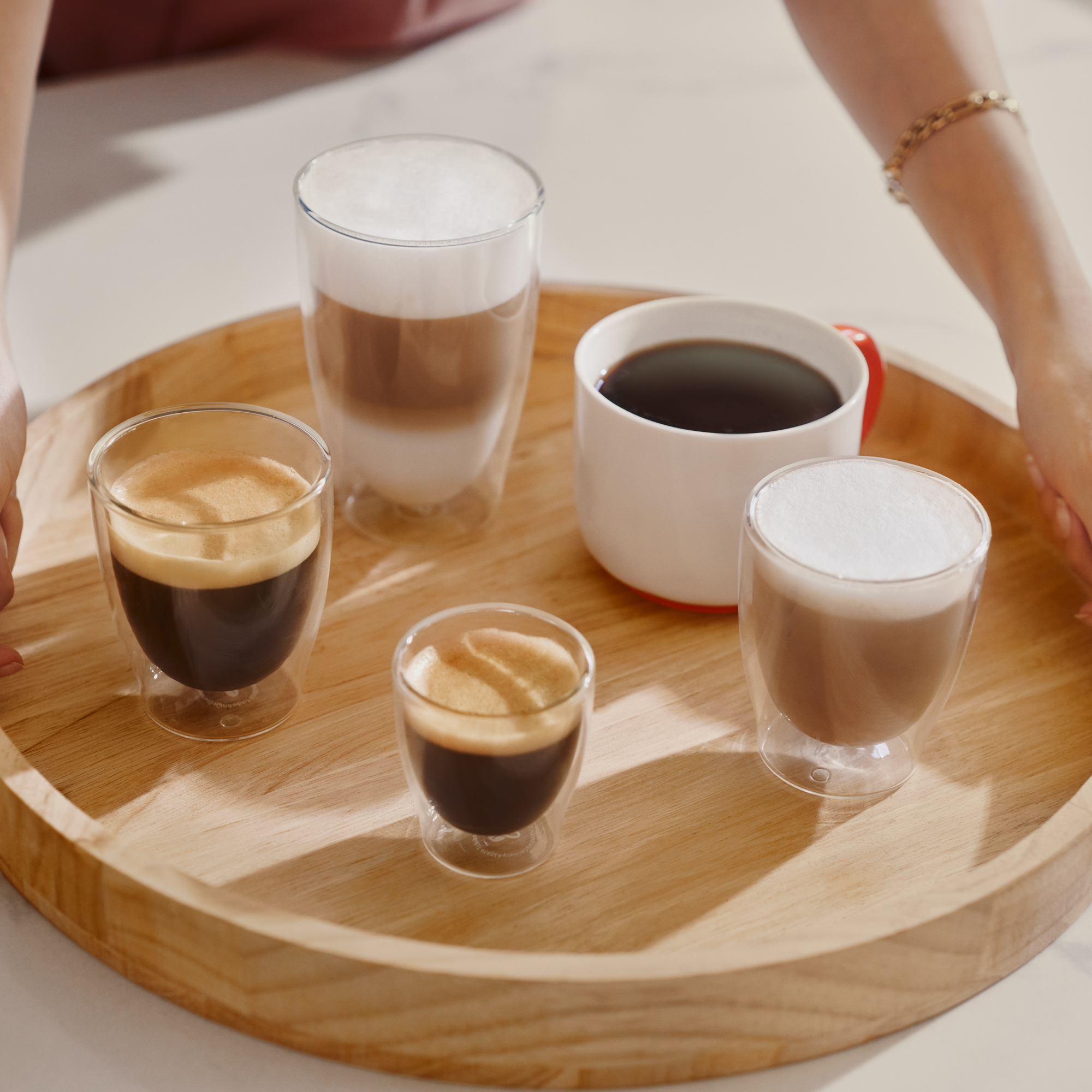 Ein hölzernes Tablett mit fünf verschiedenen Kaffeespezialitäten in Gläsern und einer Tasse wird von Händen gehalten.