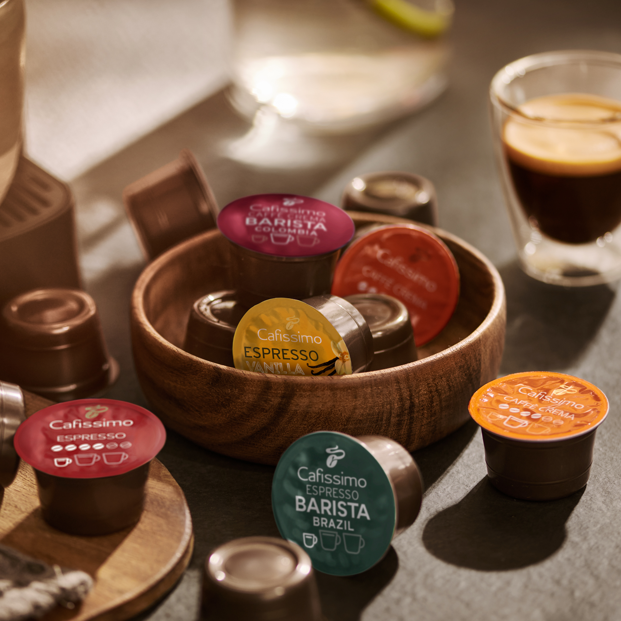 Bunte Cafissimo Kaffeekapseln in einer Holzschale auf einem Tisch mit einem Glas Espresso.