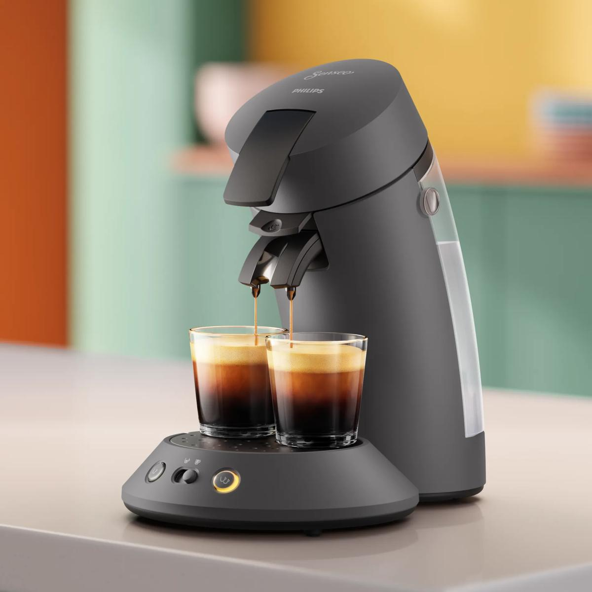 Una cafetera gris Philips Senseo prepara café en dos vasos sobre un mostrador frente a una pared colorida.