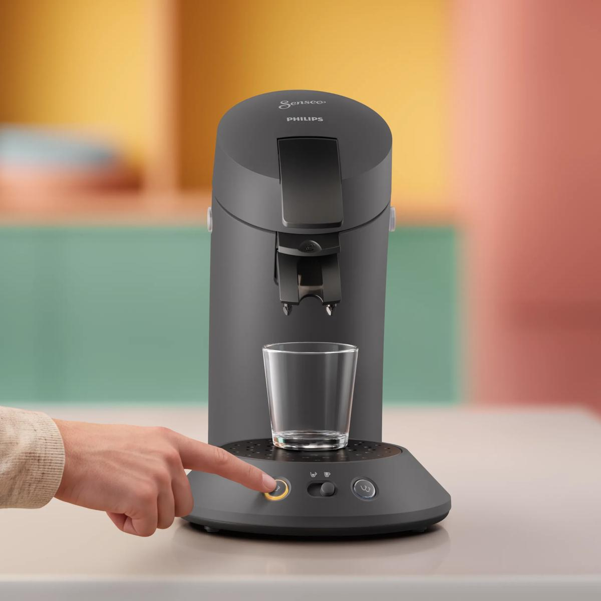 Una cafetera Philips Senseo gris sobre la mesa; una mano presiona el botón de inicio junto a un vaso.